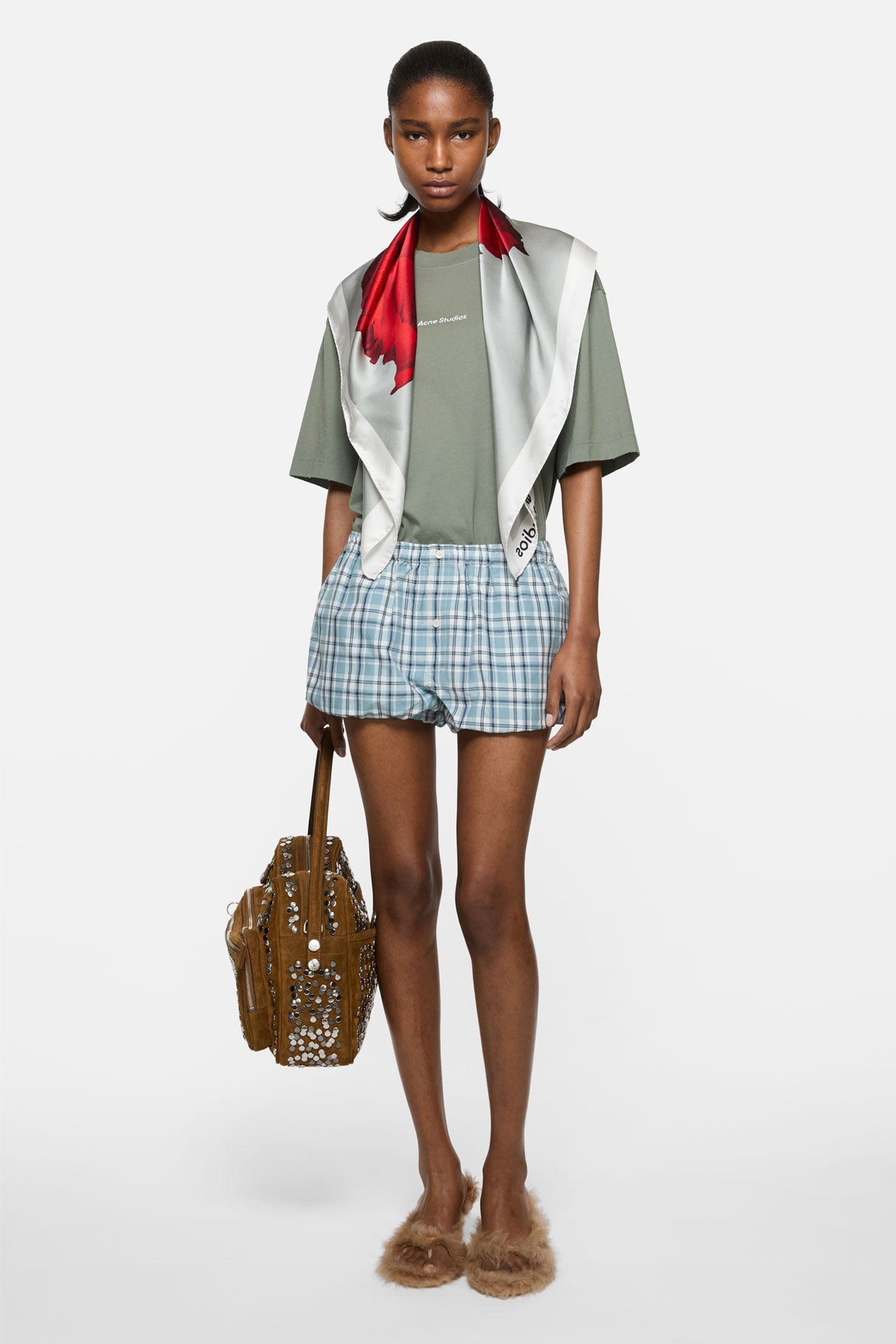 Female model wearing a sage-green t-shirt, blue-white checked shorts, a red-grey rose-printed scarf on her shoulders, holding a brown suede Camero bag.