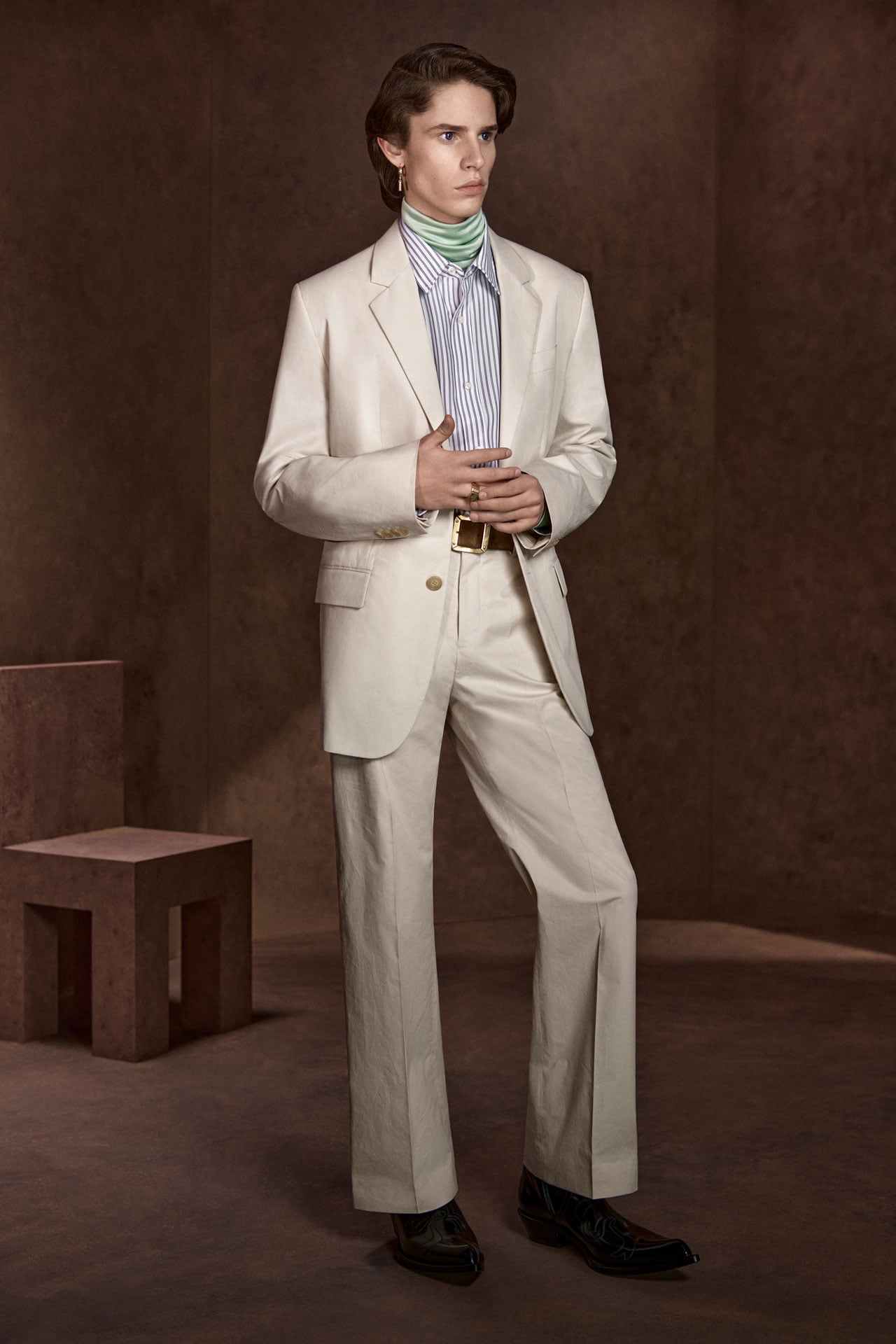 Acne Studios off-white single-breasted suit with wide-leg trousers, worn with a blue striped shirt, mint neck scarf, gold belt, and black loafers.