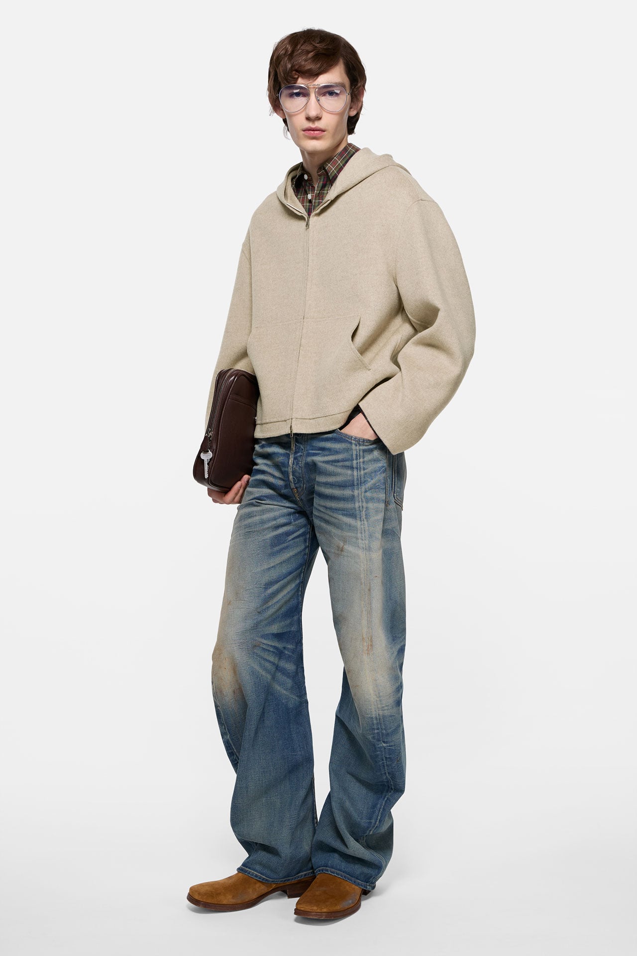 A male model wearing ripped blue jeans with a black leather belt, dark blue tanktop, brown suede cowboy boots and holding a big black leather Camero bag on his shoulder.