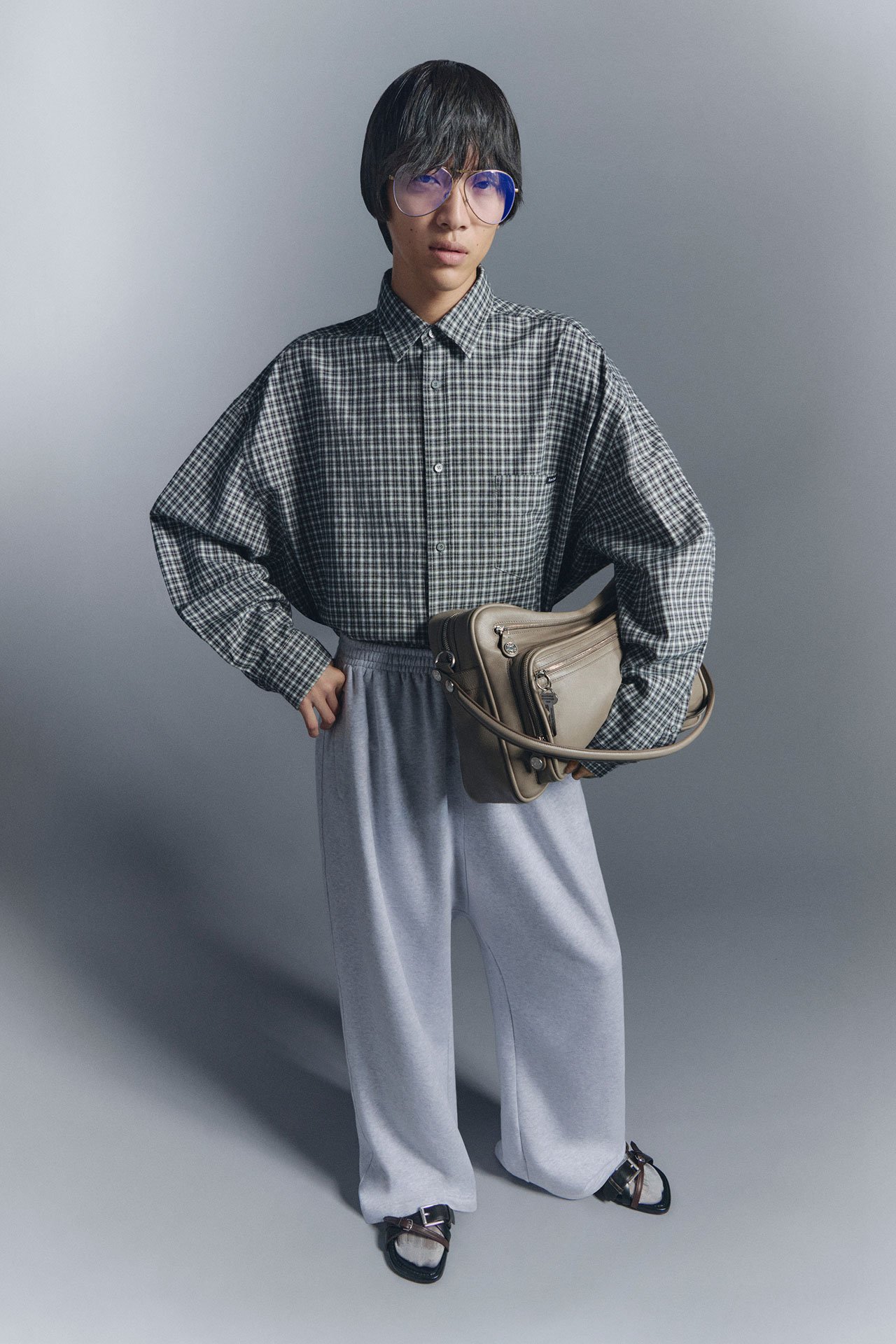 A male model wearing checked shirt, grey sweatpants, glasses and slippers, holding a taupe leather Camero bag.