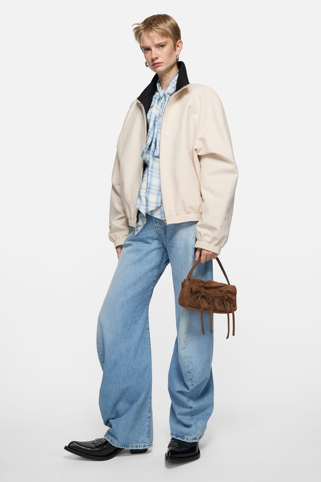 A female model wearing a white jacket, blue jeans, light blouse and holding an Acne Studios brown Multipocket bag.