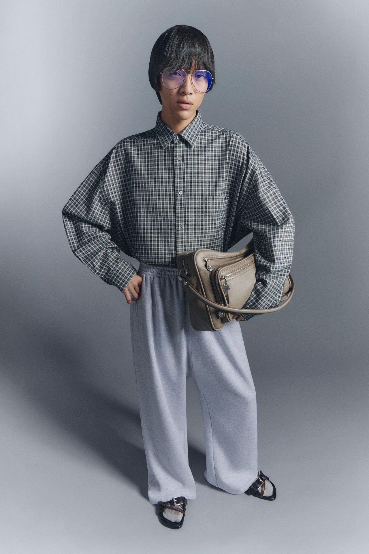 In front of a grey background a man wearing glasses, grey pants, slippers and checked shirt, holding a taupe Camero bag.