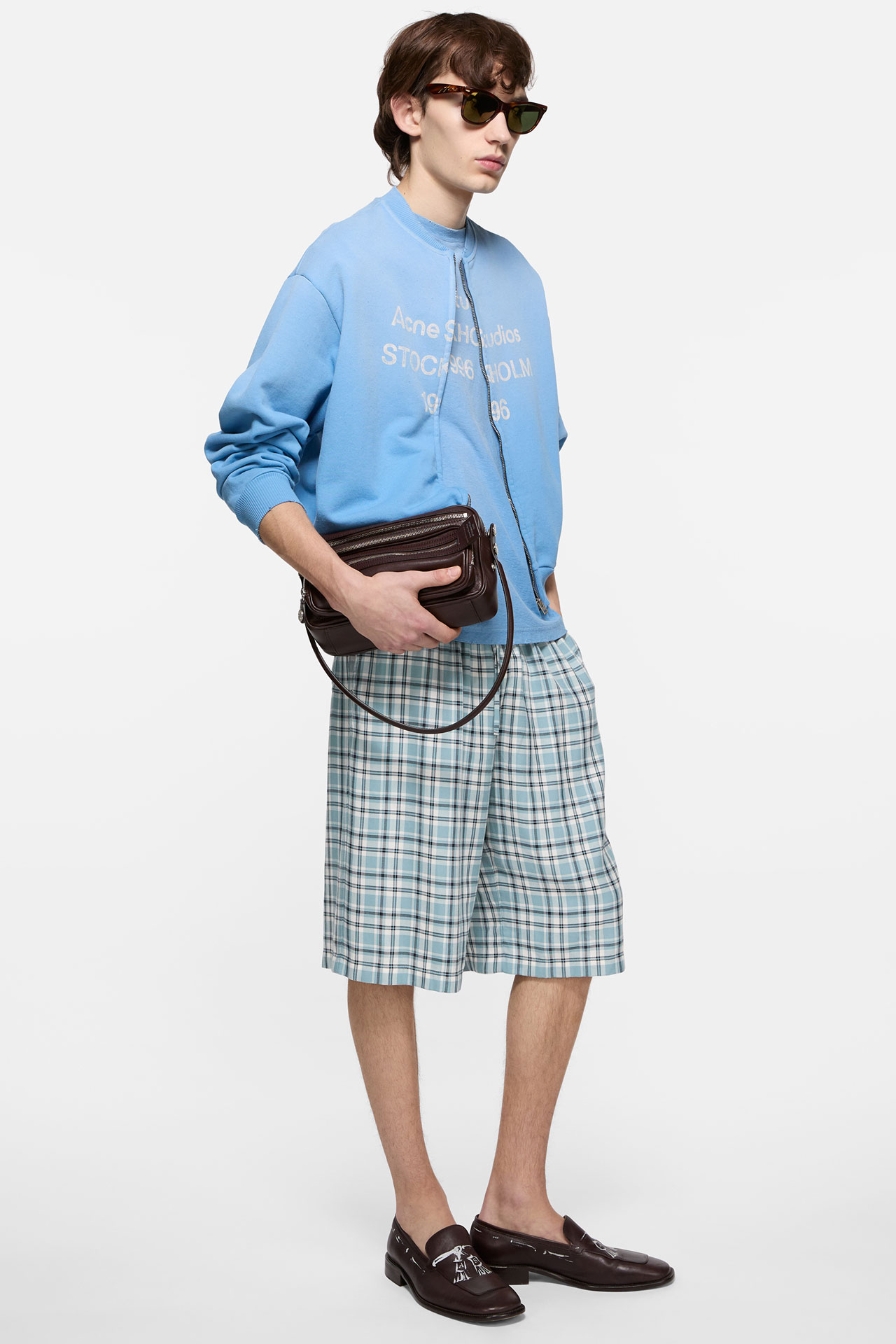 Male model wearing and Acne Studios cornflower blue zipper, matching t-shirt, blue checked shorts and holding a brown leather Camero bag.