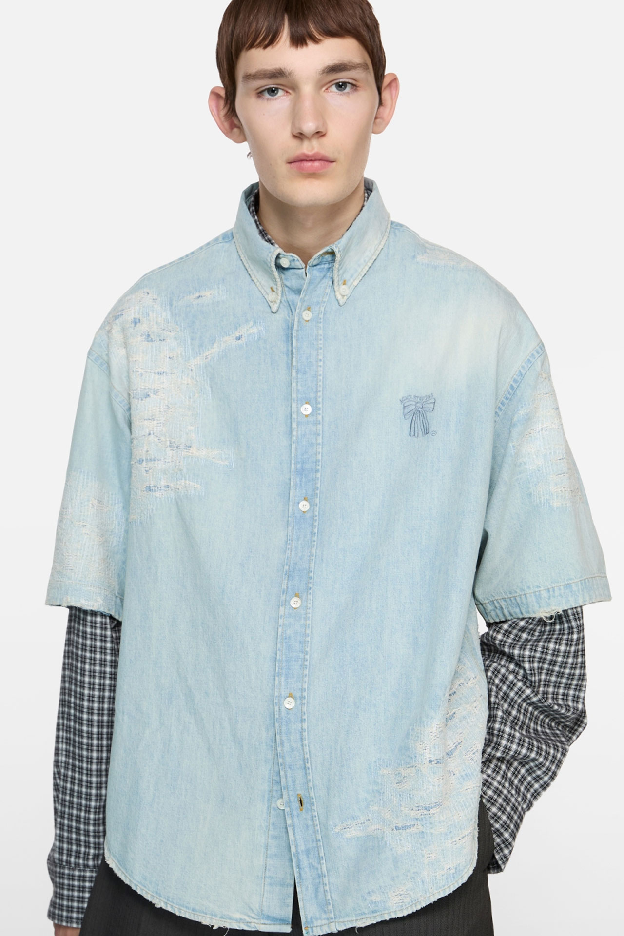 Male model wearing and Acne Studios light blue denim shirt, dark checked long-sleeve t-shirt under, and black pants.