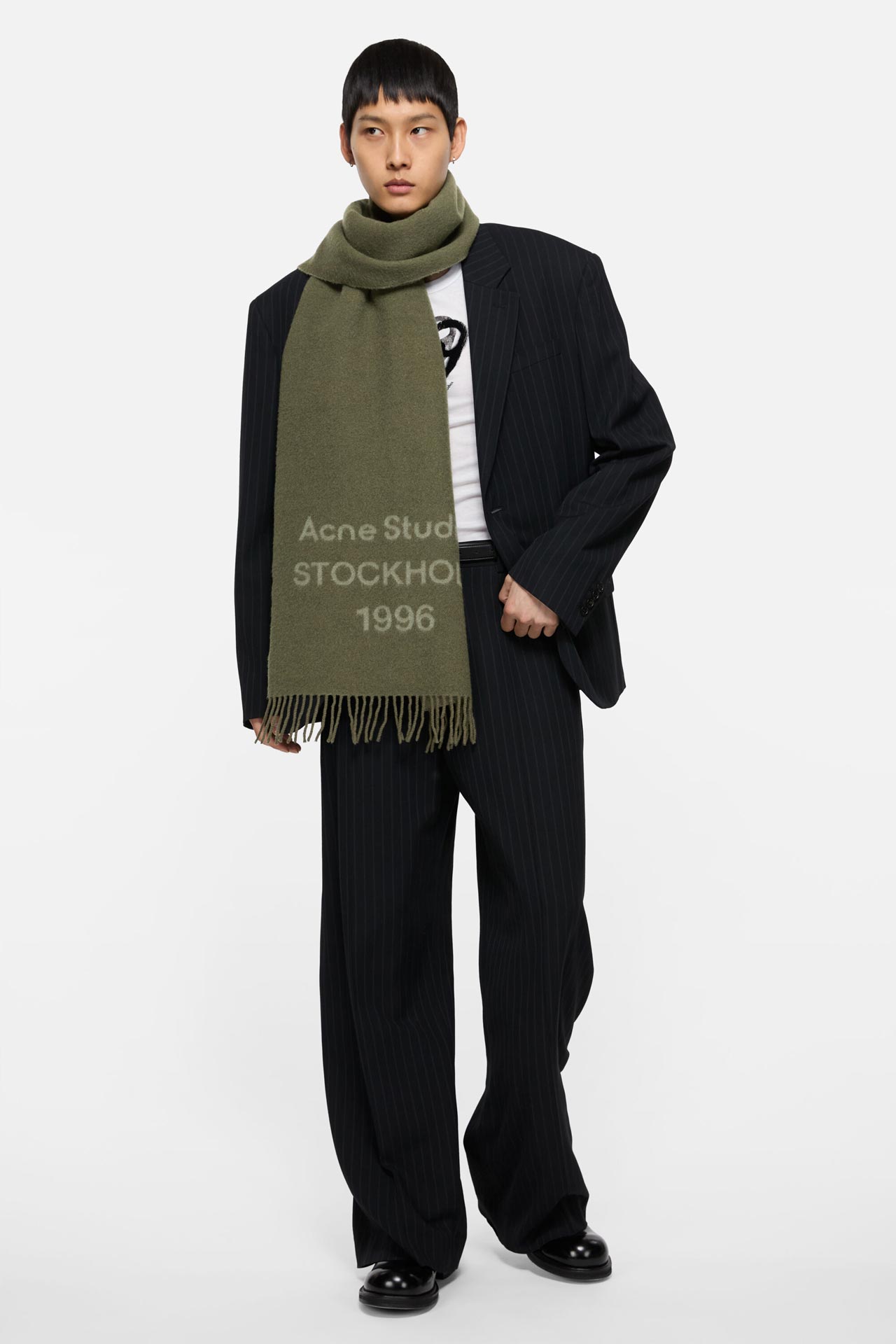 Male model wearing an Acne Studios 1996 logo scarf in olive green, and matching black suit jacket and pants with stripes.