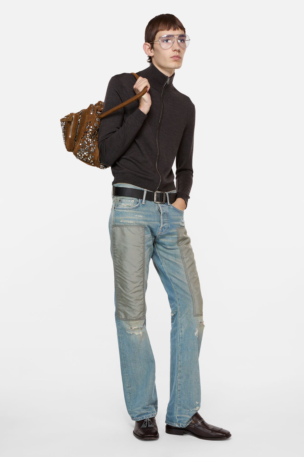 A male model wearing a grey cashmere zipper cardigan, blue jeans, brown leather loafers and holding a brown suede Camero bag.