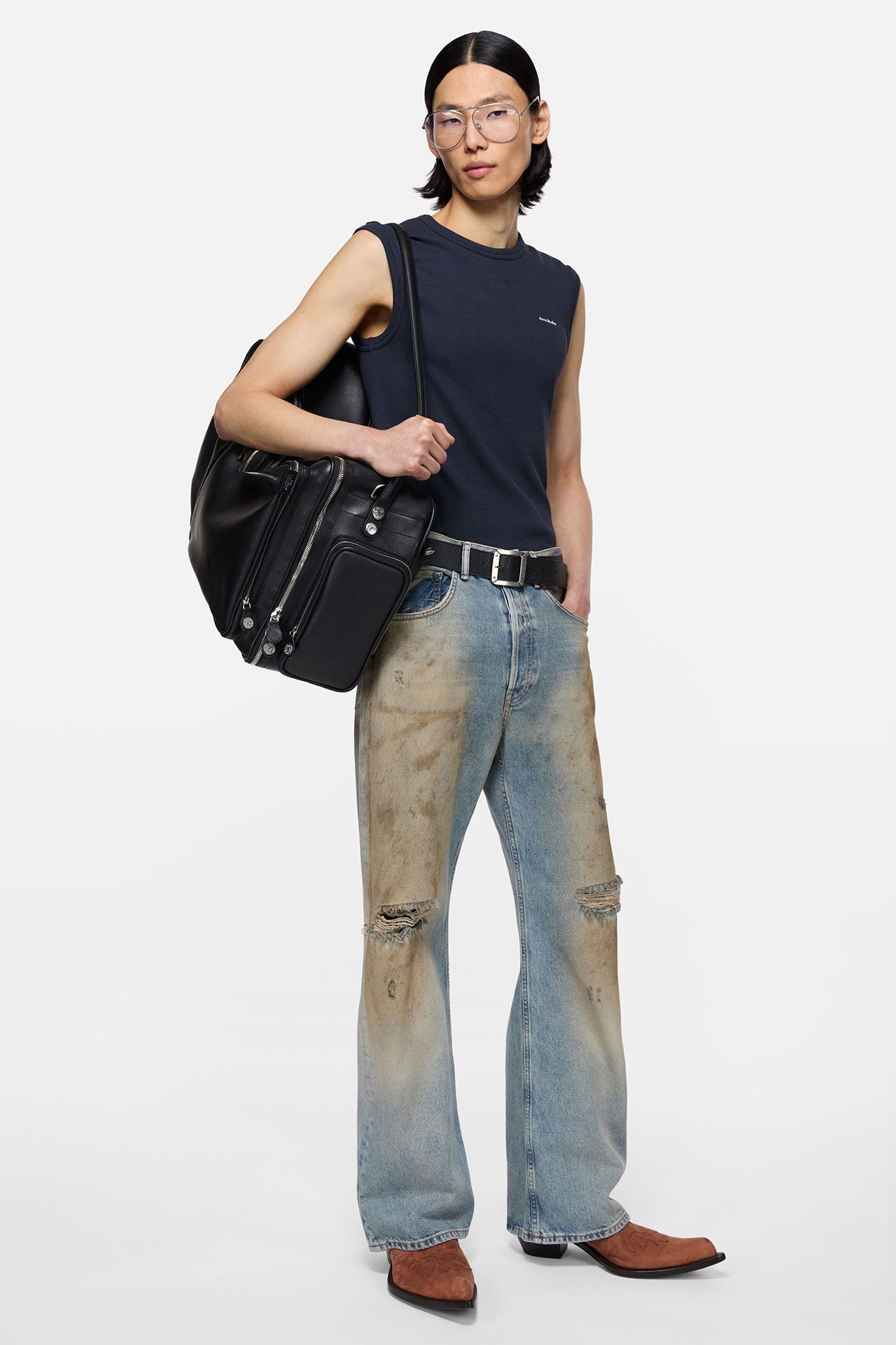 A male model wearing ripped blue jeans with a black leather belt, dark blue tanktop, brown suede cowboy boots and holding a big black leather Camero bag on his shoulder.