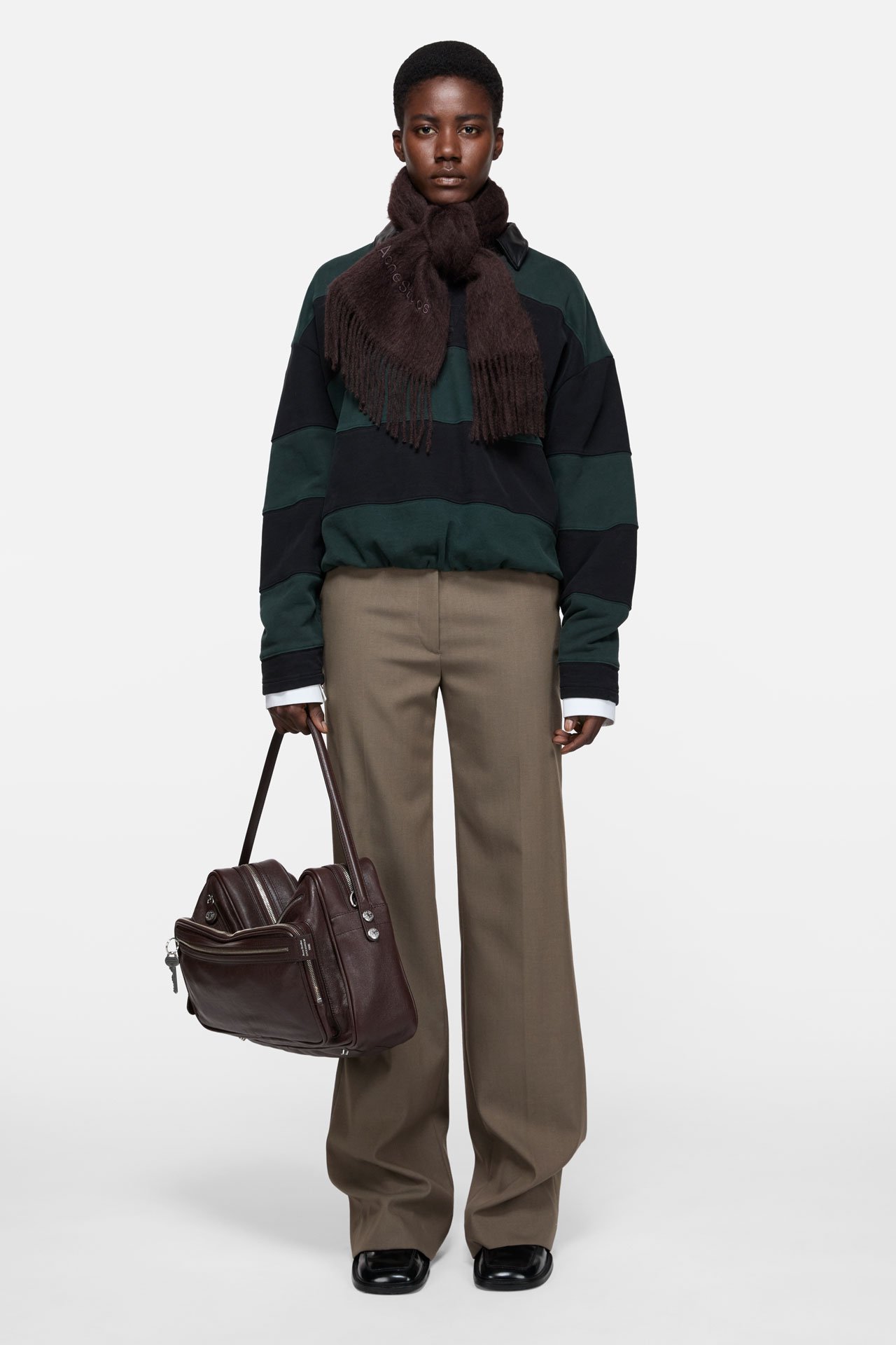 A female model wearing Acne Studios brown wool logo scarf, green and black striped long-sleeve shirt, beige pants, holding a big brown leather Camero bag.