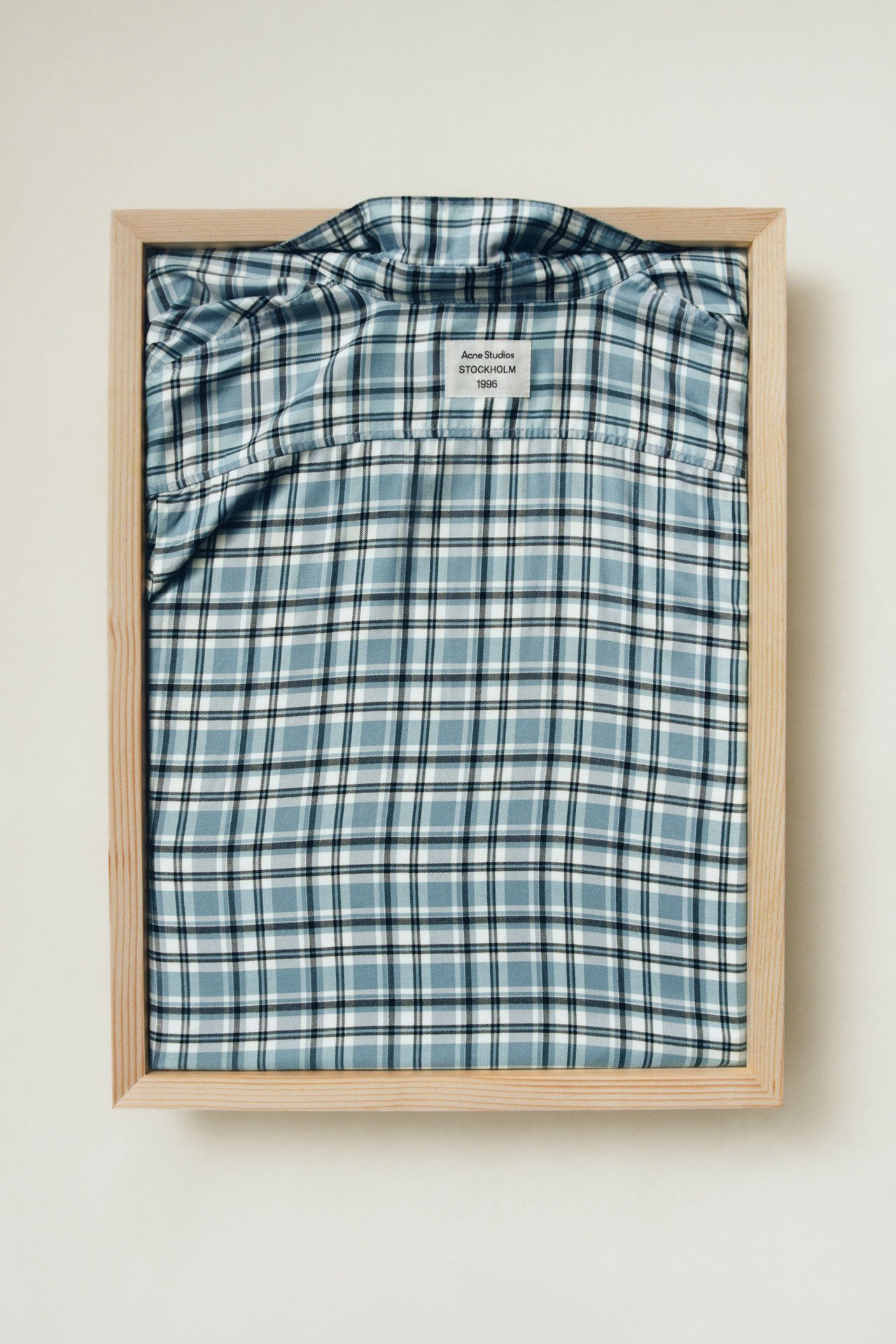 An Acne Studios blue checked shirt folded in a light wooden tray.