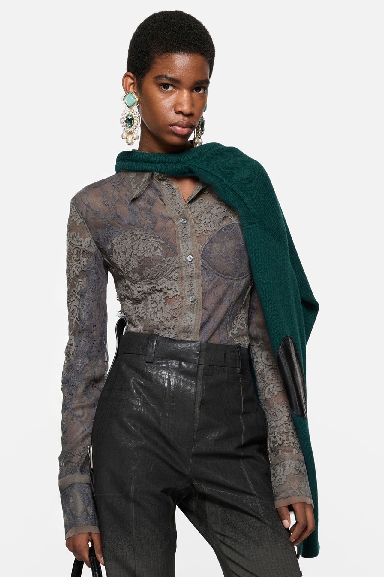 A female model wearing Acne Studios dark grey shirt with lace details, black leather pants, green knitted sweater on her shoulder and a pair of bold tile-colored earrings.