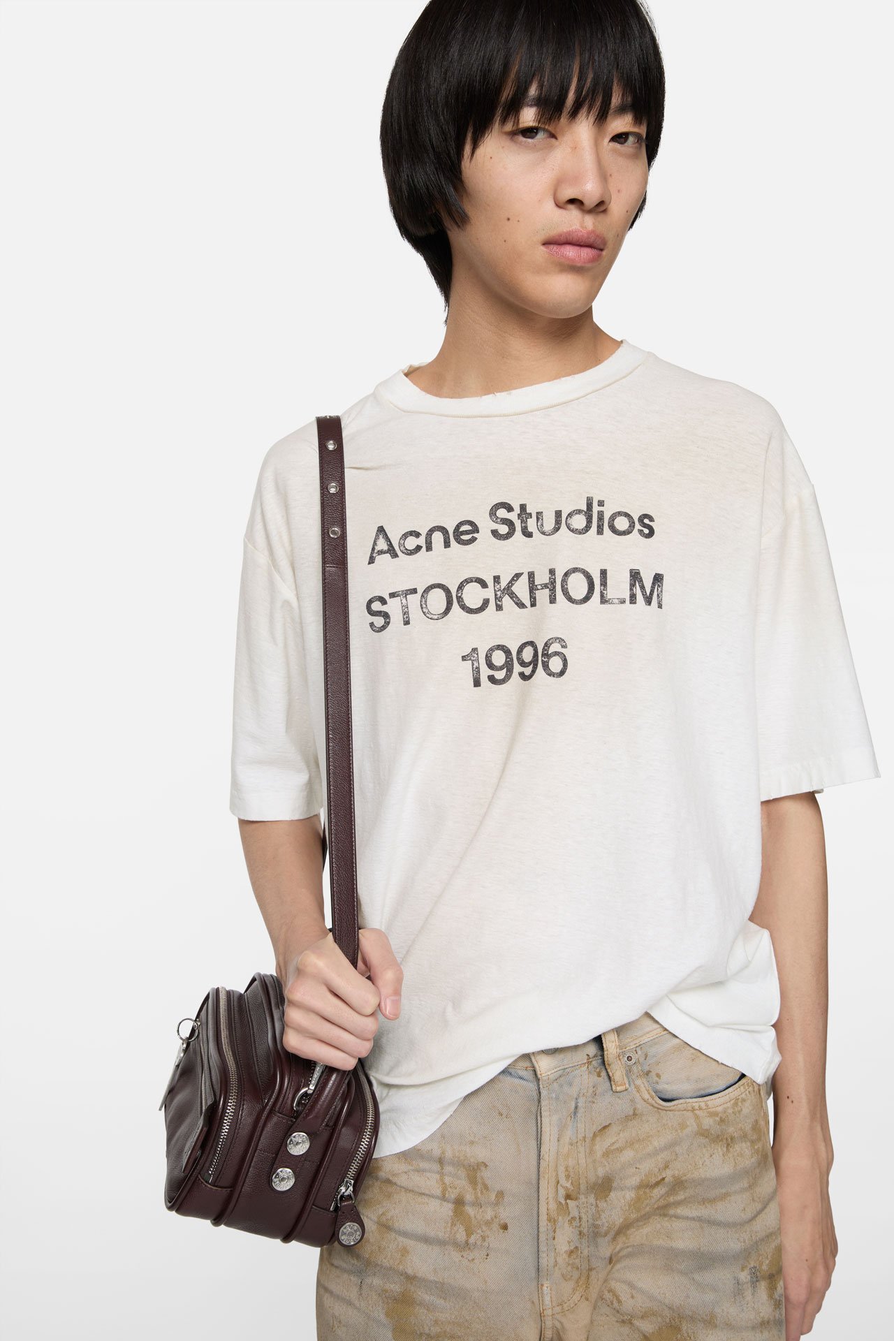 A male model wearing an Acne Studios 1996 logo white t-shirt, beige jeans and holding a brown leather Camero bag.