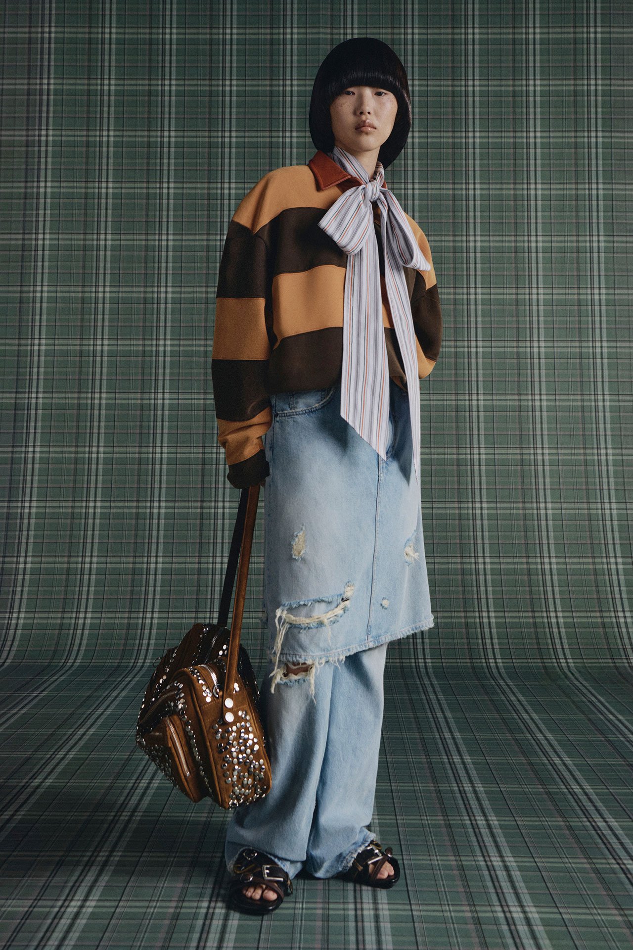 Female model with glasses is wearing Acne Studios SS26 new arrivals brown striped long-sleeve t-shirt, blue jeans, slippers, a light blue scarf tied in a bow shape, holding a big brown Camero bag, in front of a green checked background.