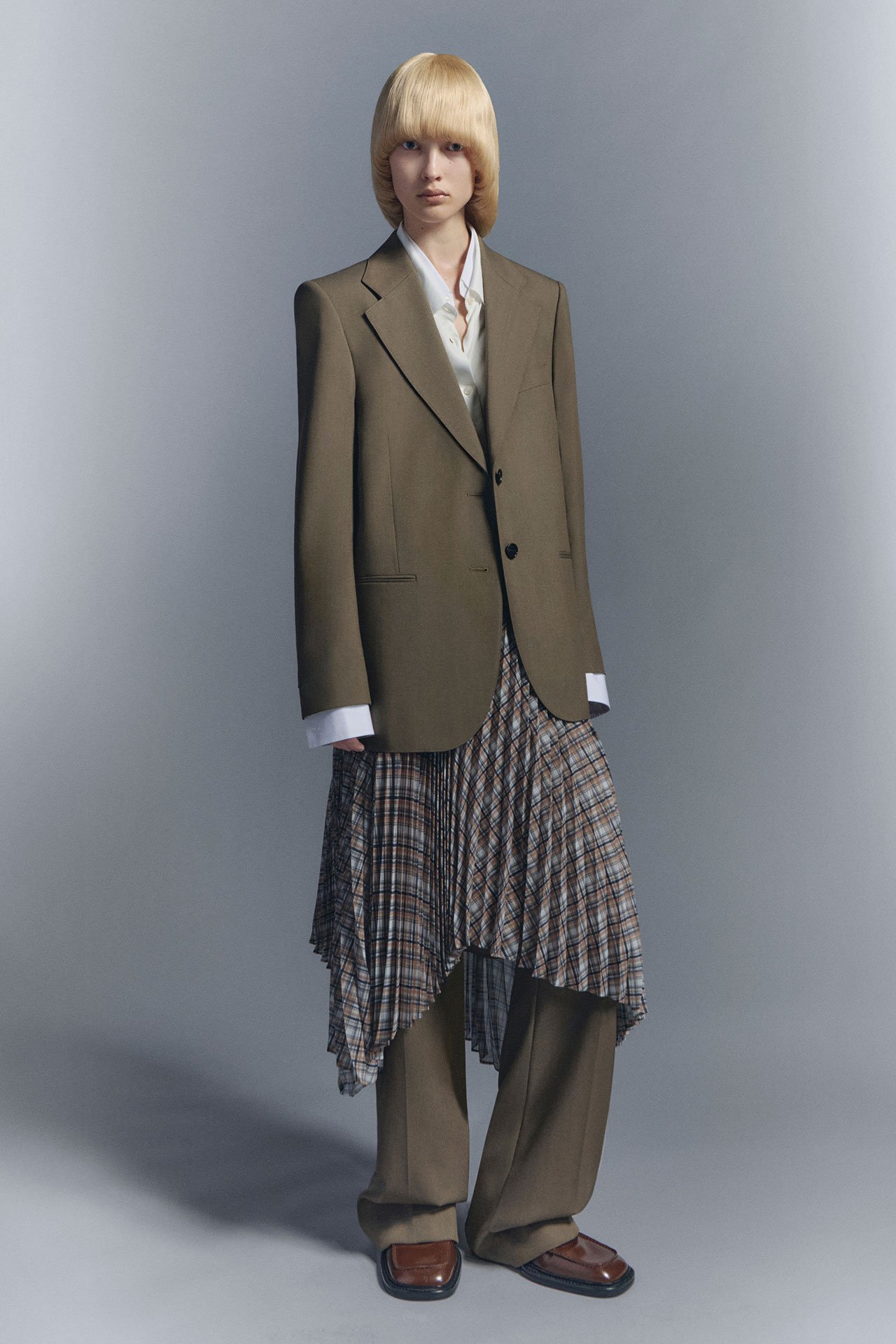 Female model wearing taupe tailored suite jacket, matching tailored pants, checked skirt and white shirt in front of a creative grey background.