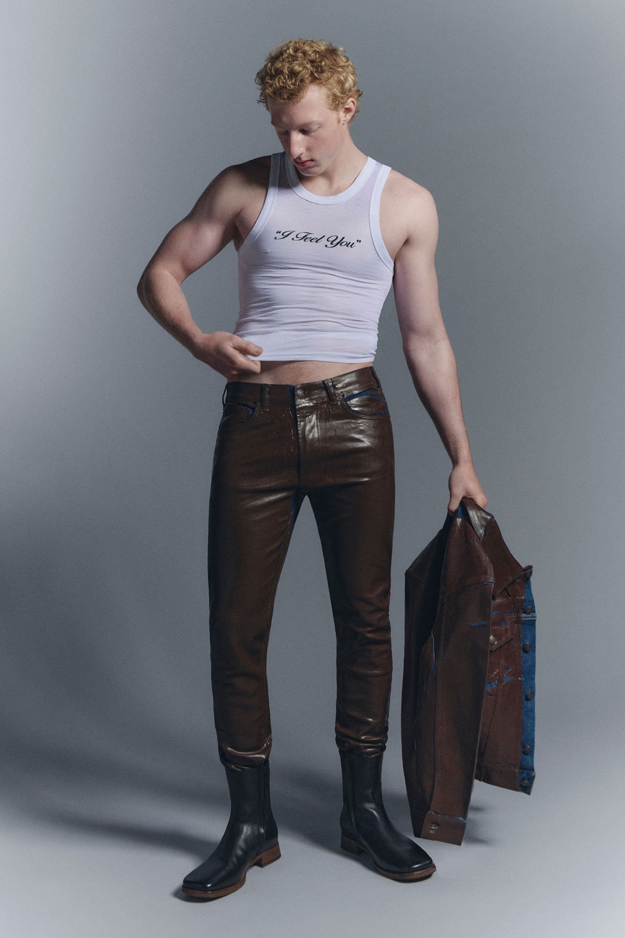 A male model wearing a white topless shirt, brown leather pants, black leather boots and holding a brown leather jacket.