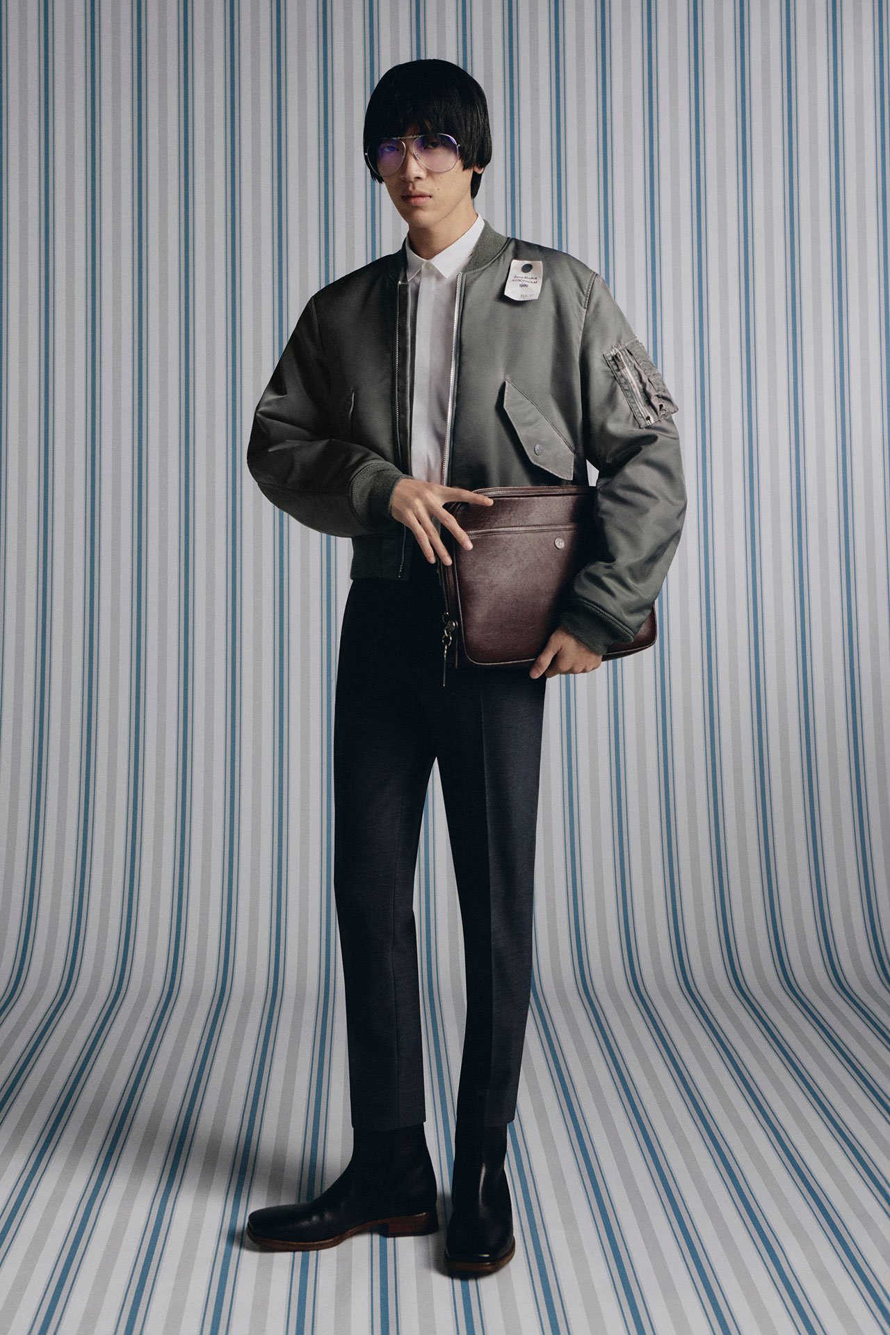 A male model wearing green bomber jacket, tight black pants, white shirt, holding a brown leather laptop case in his hands in front of a striped background.