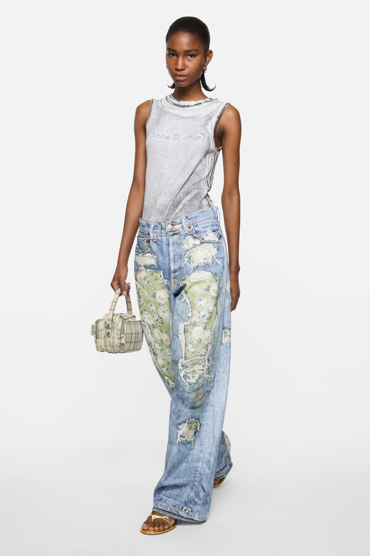 A female model wearing Acne Studios green-blue TLO jeans with rose patterns, a grey tanktop and holding a green leather Camero bag.