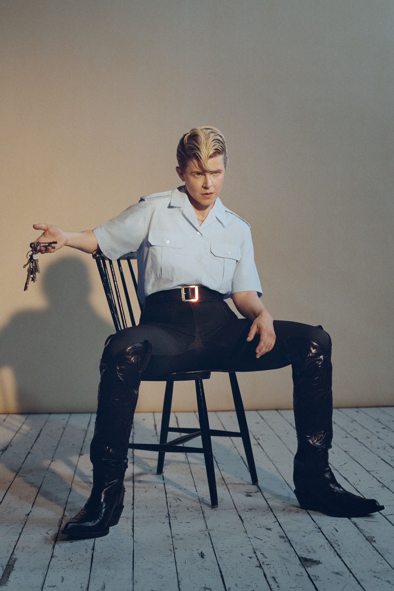 Artist Robyn posing in black jeans and white-beige short-sleeve shirt on a chair, holding a keychain.
