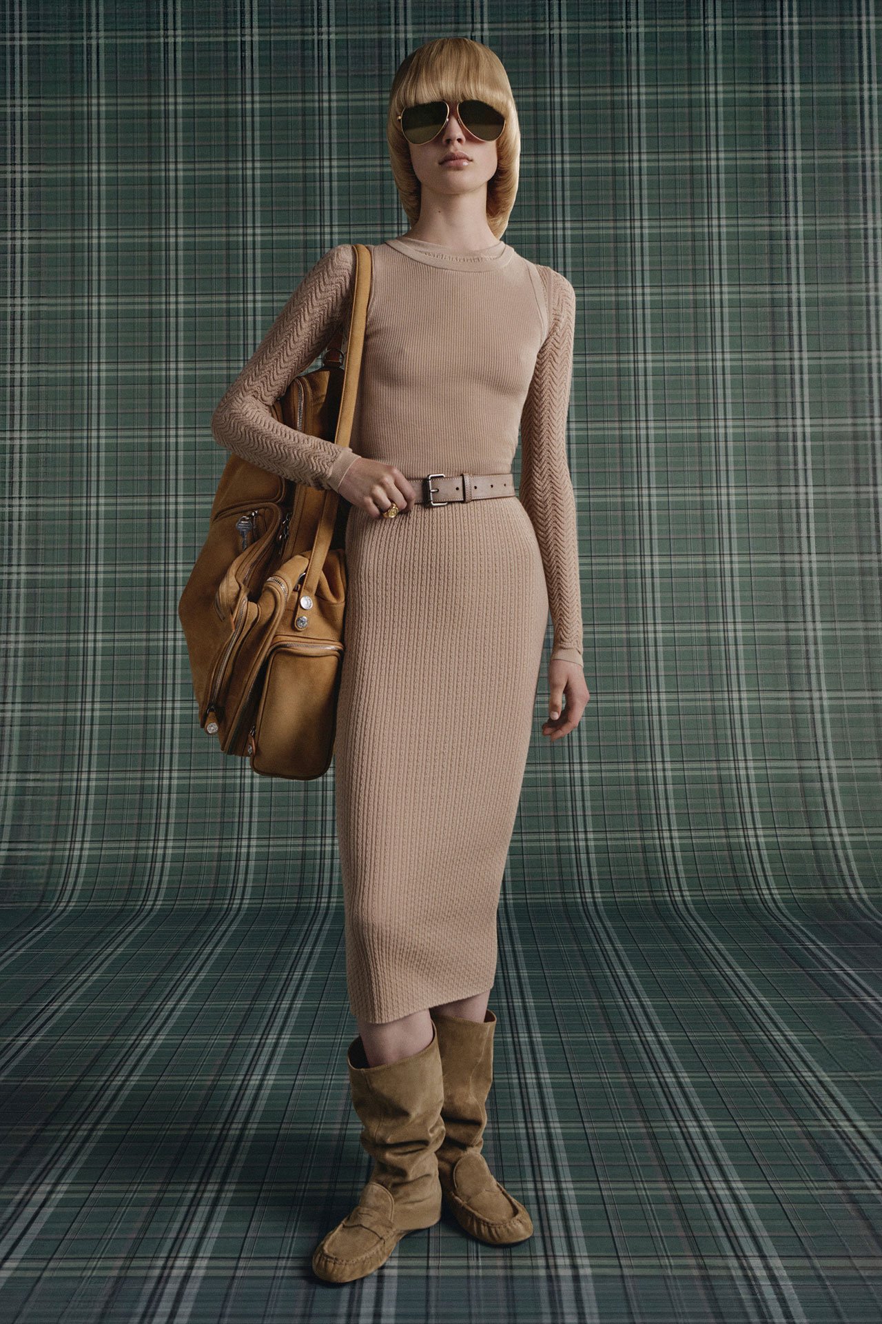 Female model wearing a beige long fitted dress with a matching belt, mirrored-sunglasses, beige suede boots and holding a big beige suede Camero Hotel bag.