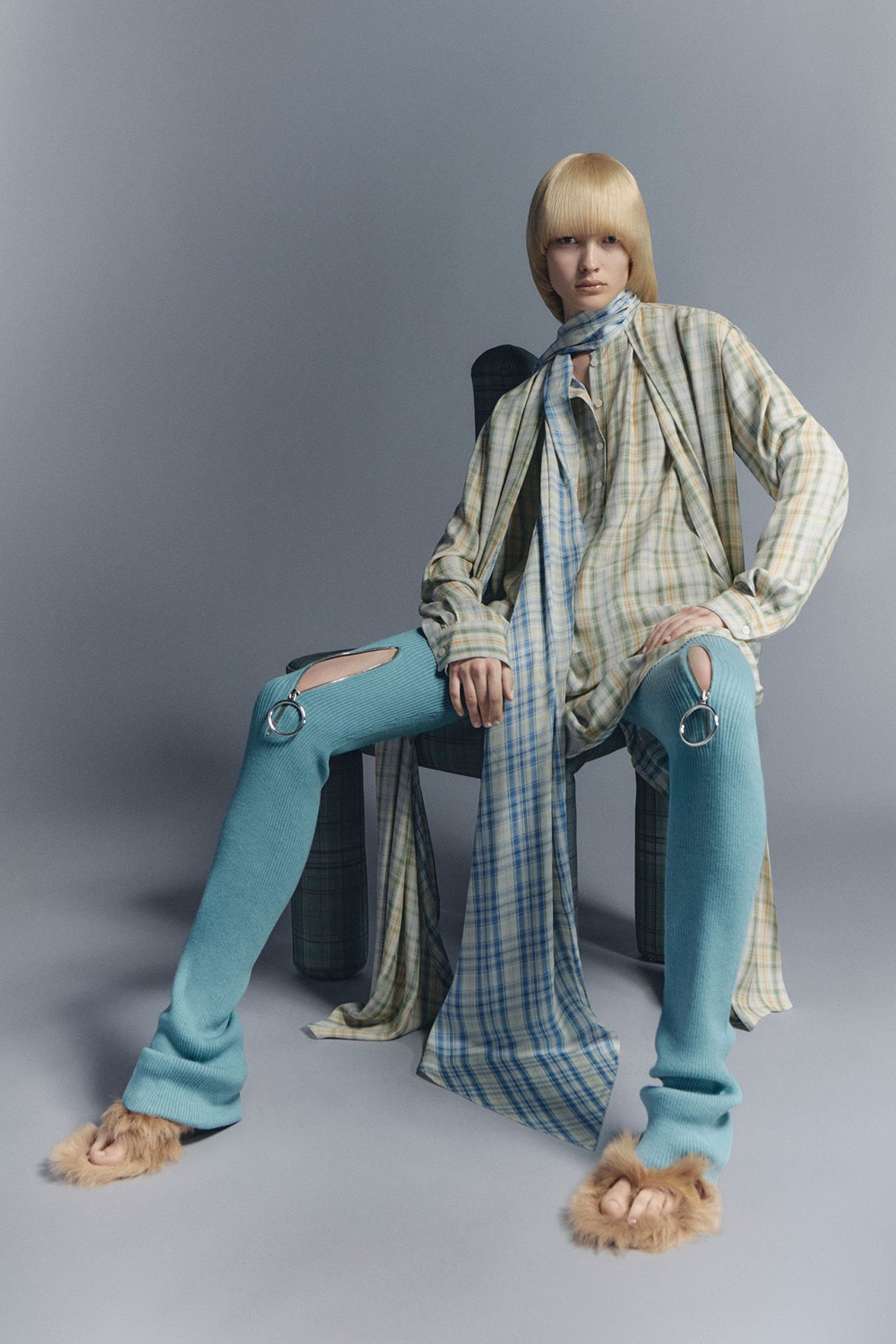 SS26 Acne Studios Womens Fluid Silhouettes - female model wearing blue pants, blue-yellow checked shirt with scarf and taupe fluffy slippers, sitting on a chair.