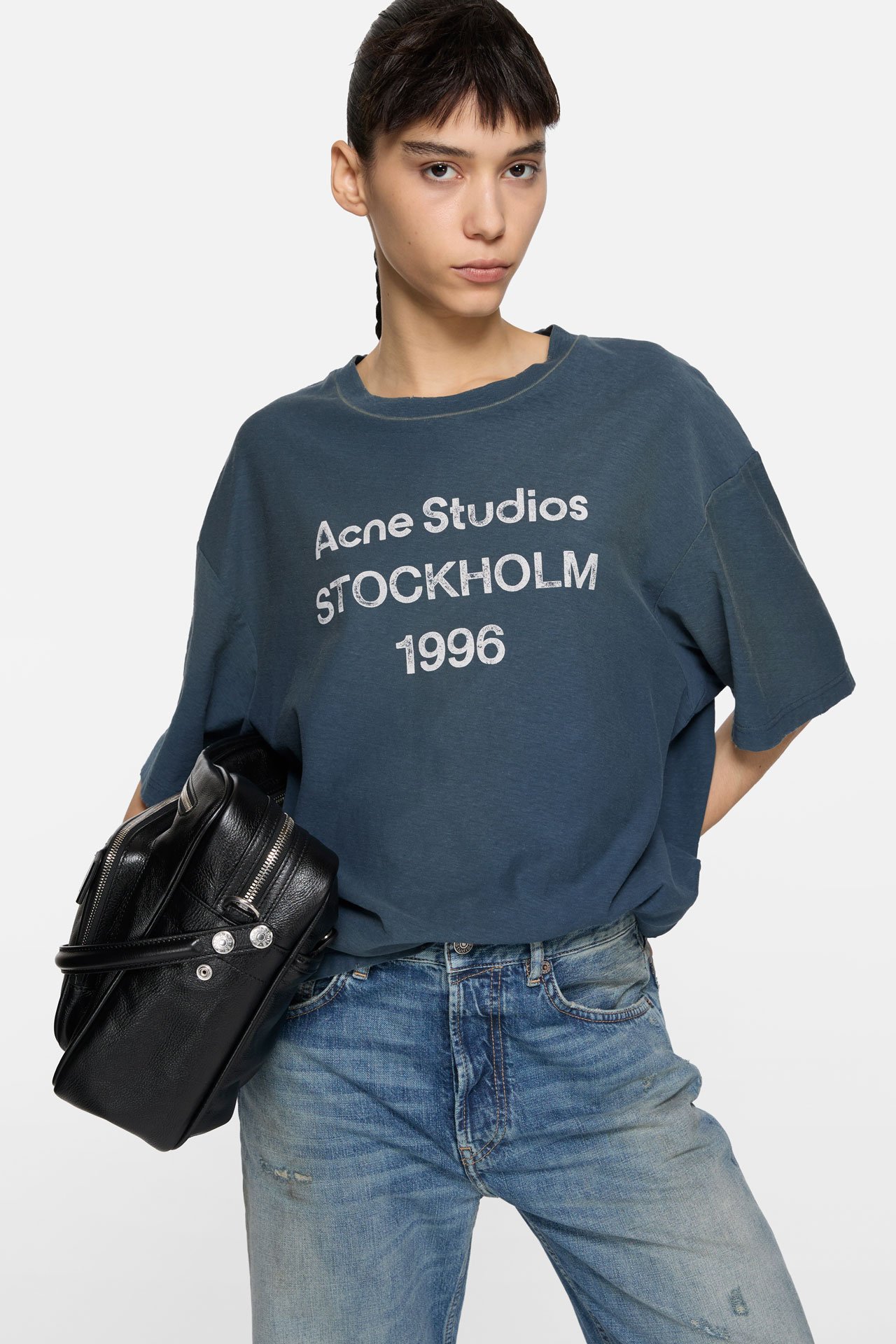 A female model wearing an Acne Studios 1996 logo dark blue t-shirt and wearing jeans, holding a black leather Camero bag.