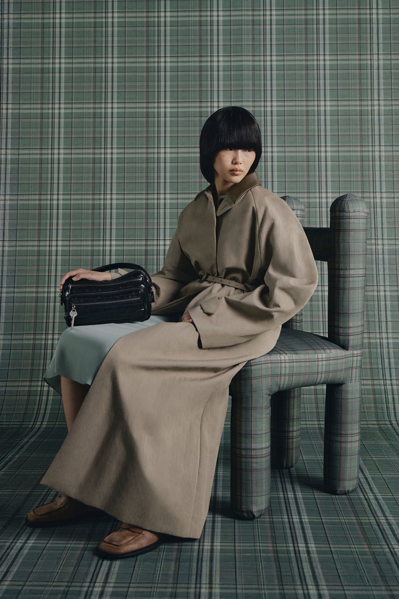 Female model wearing a long taupe trench coat and brown loafers sitting on a chair, holding a black leather Camero bag in front of a green checked background.