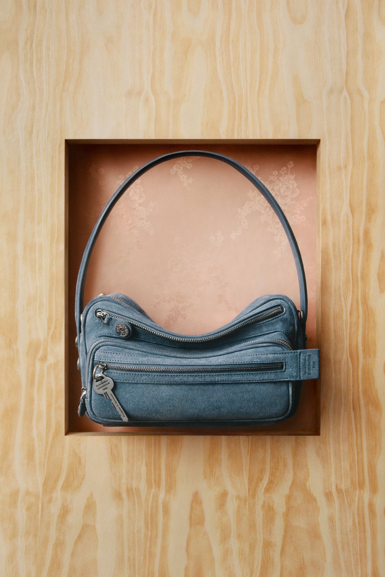 An Acne Studios blue suede Camero bag presented on a light wooden panel.
