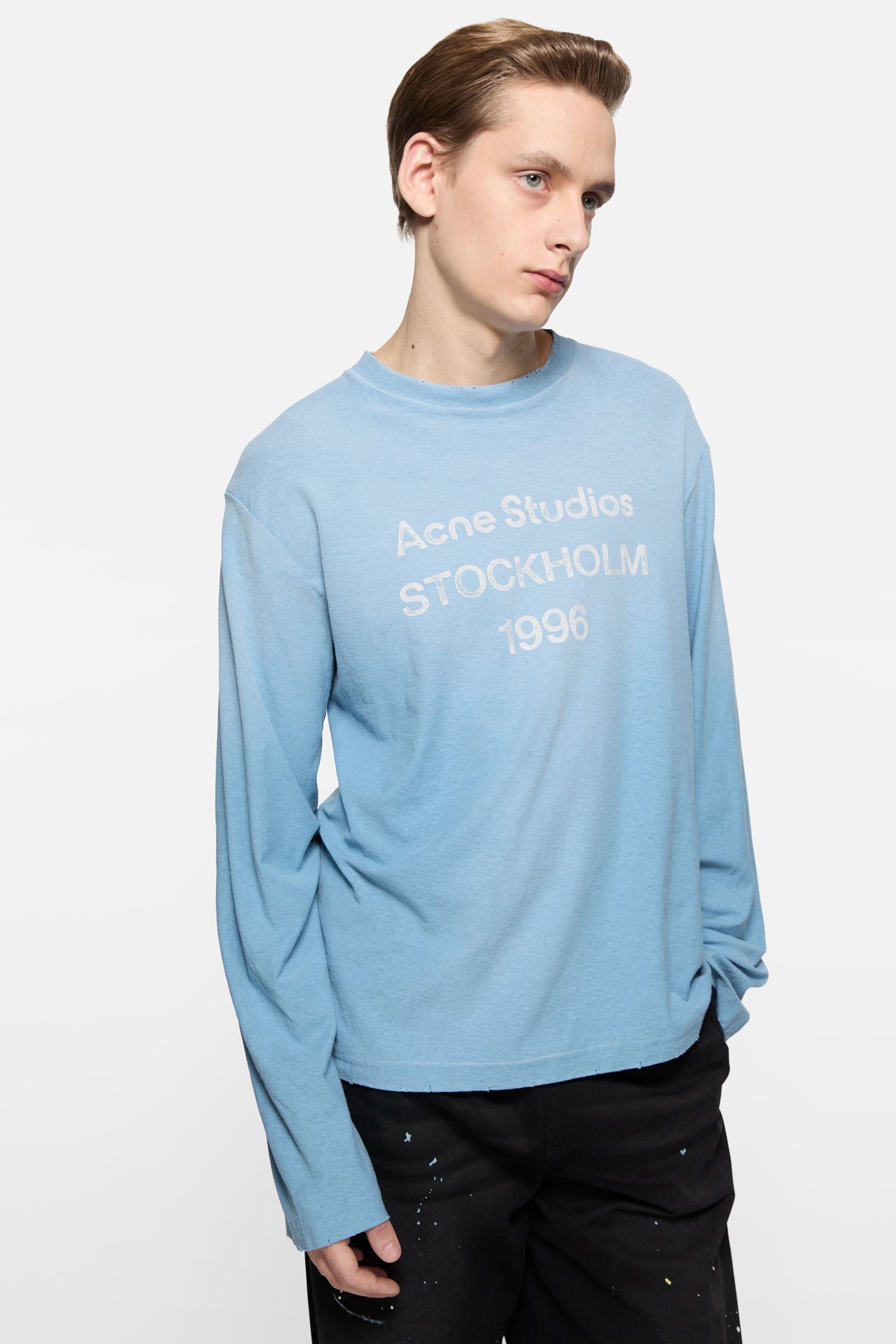 A male model wearing an Acne Studios 1996 logo cornflower blue t-shirt and black pants.