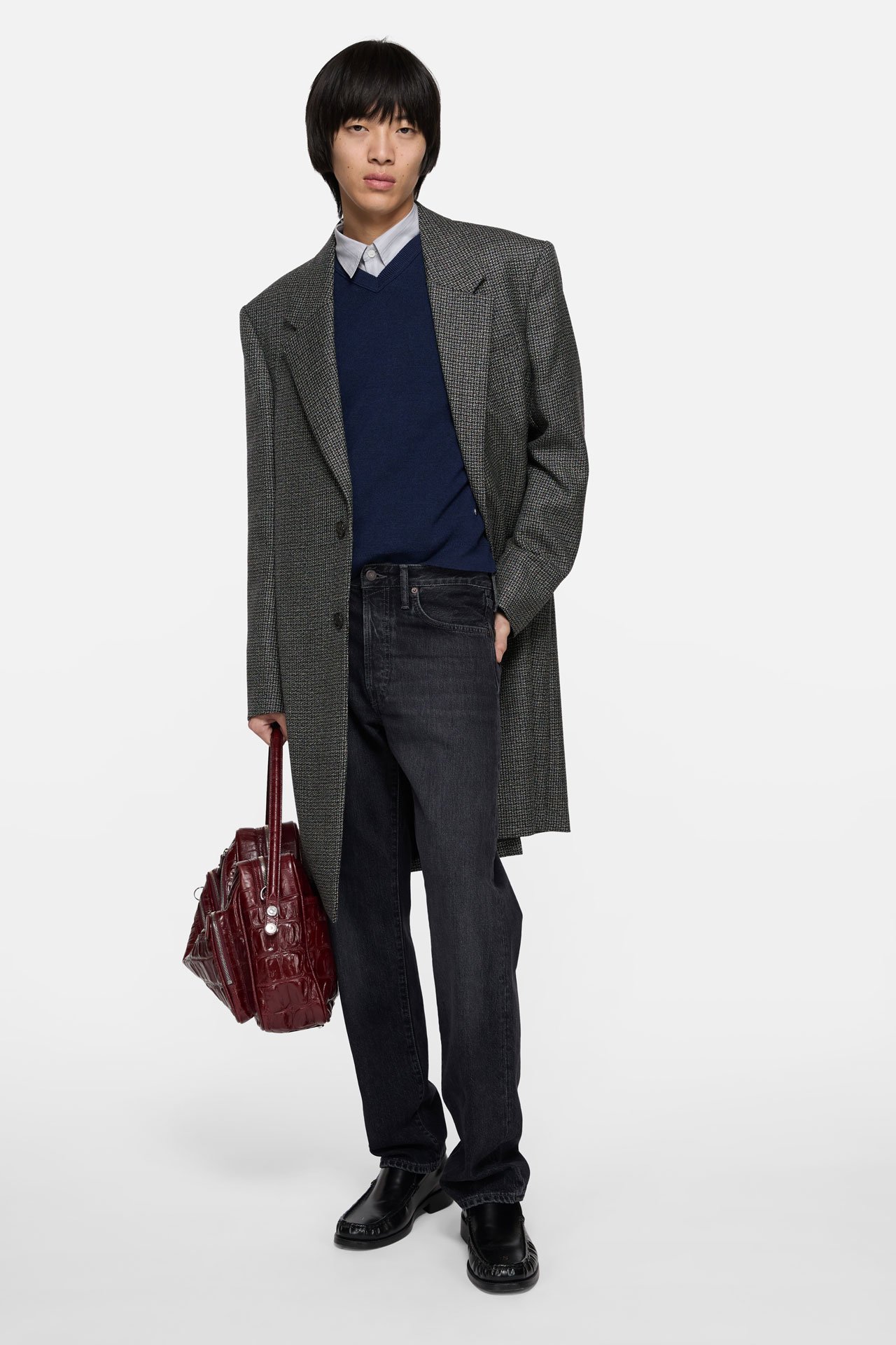 A male model wearing Acne Studios long grey tailored suit jacket, blue knitted sweater, black washed jeans and black leather loafers, holding a brown leather Camero bag.