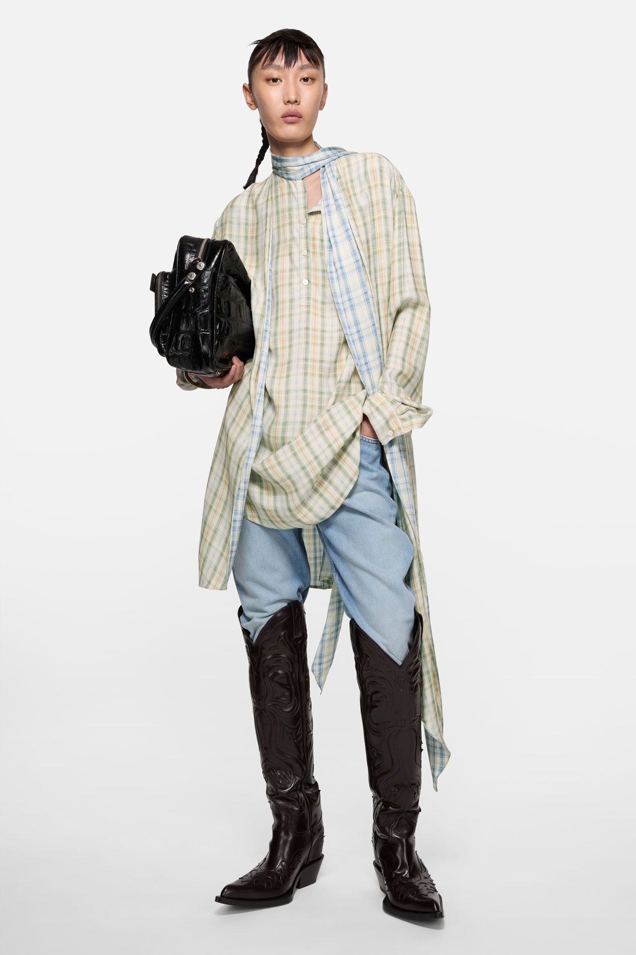 Female model wearing a light green-blue checked dress and blue pants, long brown leather cowboy boots and holding a black leather Camero bag.