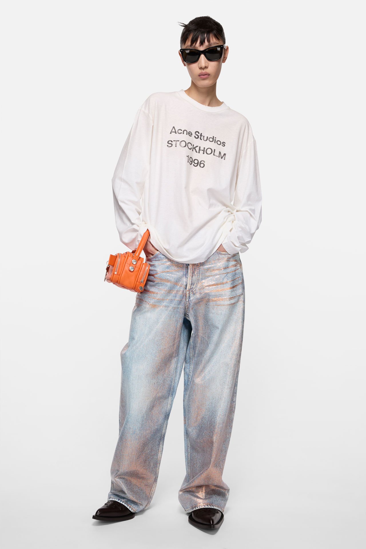 A female model wearing Acne Studios loose glitter jeans - 1981, white long-sleeve logo t-shirt and holding an orange leather Camero bag.