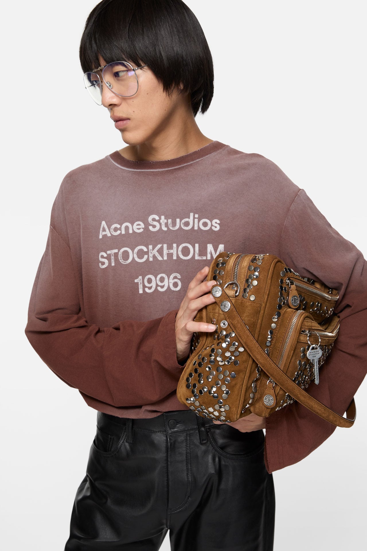 A male model wearing an Acne Studios 1996 logo brown long-sleeve t-shirt, holding a brown suede Camero bag.