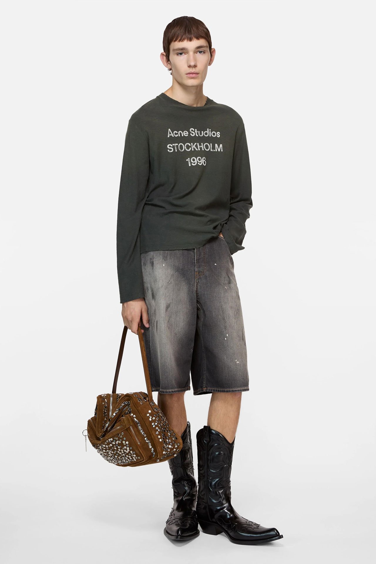 Male model showing Acne Studios new arrivals, wearing a long grey tailored suit jacket, pink hoodie, black leather pants, loafers and holding a colorful bag.