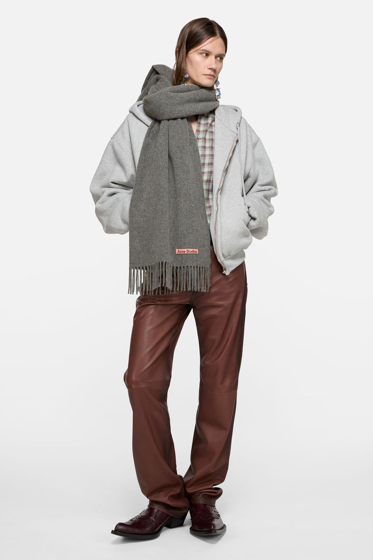 A female model wearing a Canada fringe grey wool scarf, thick grey sweathers, bold silver earrings and brown leather pants.