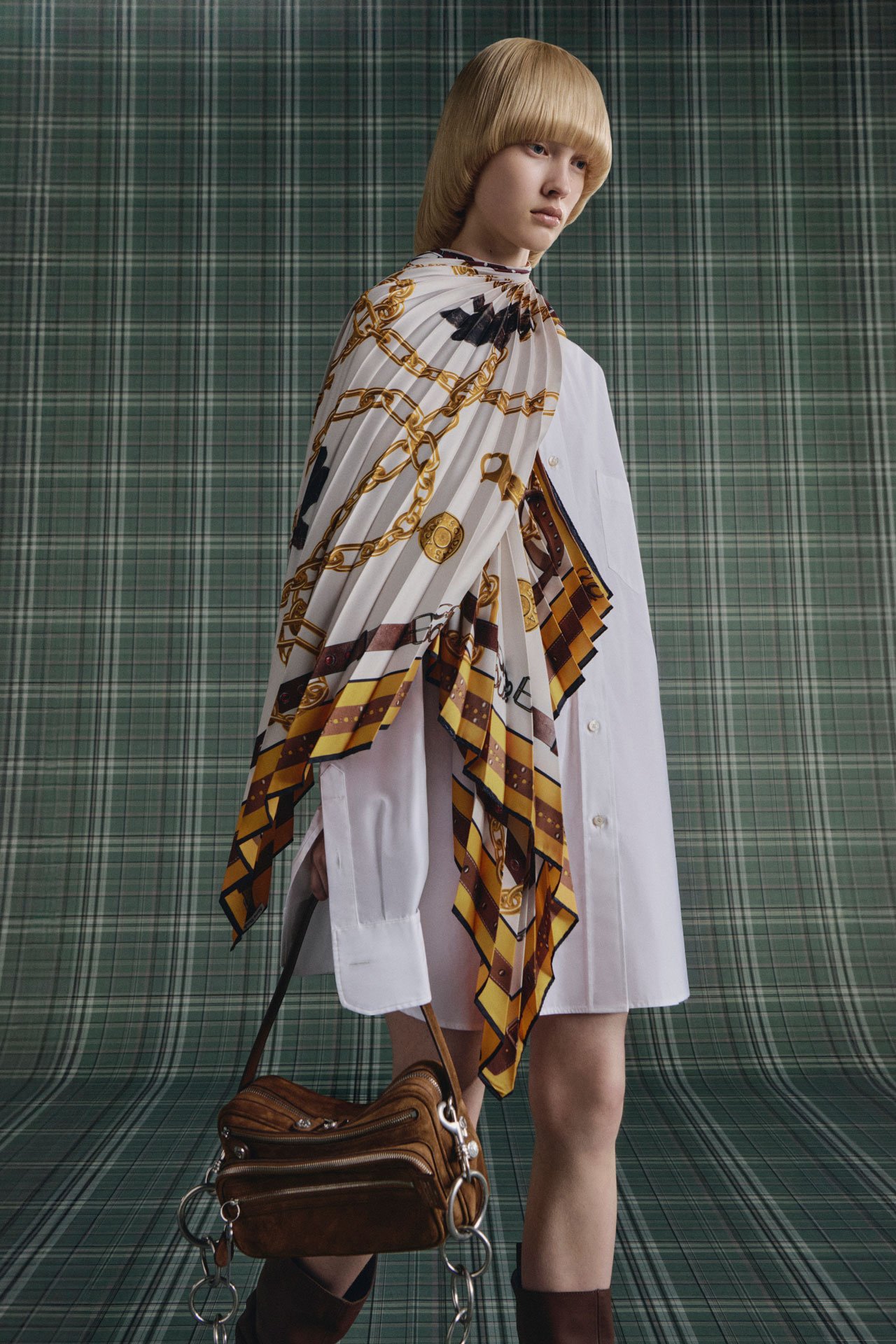 Blond woman in Acne Studios SS26 new arrivals short white dress with a white-gold scarf, holding a brown faux leather Camero bag.