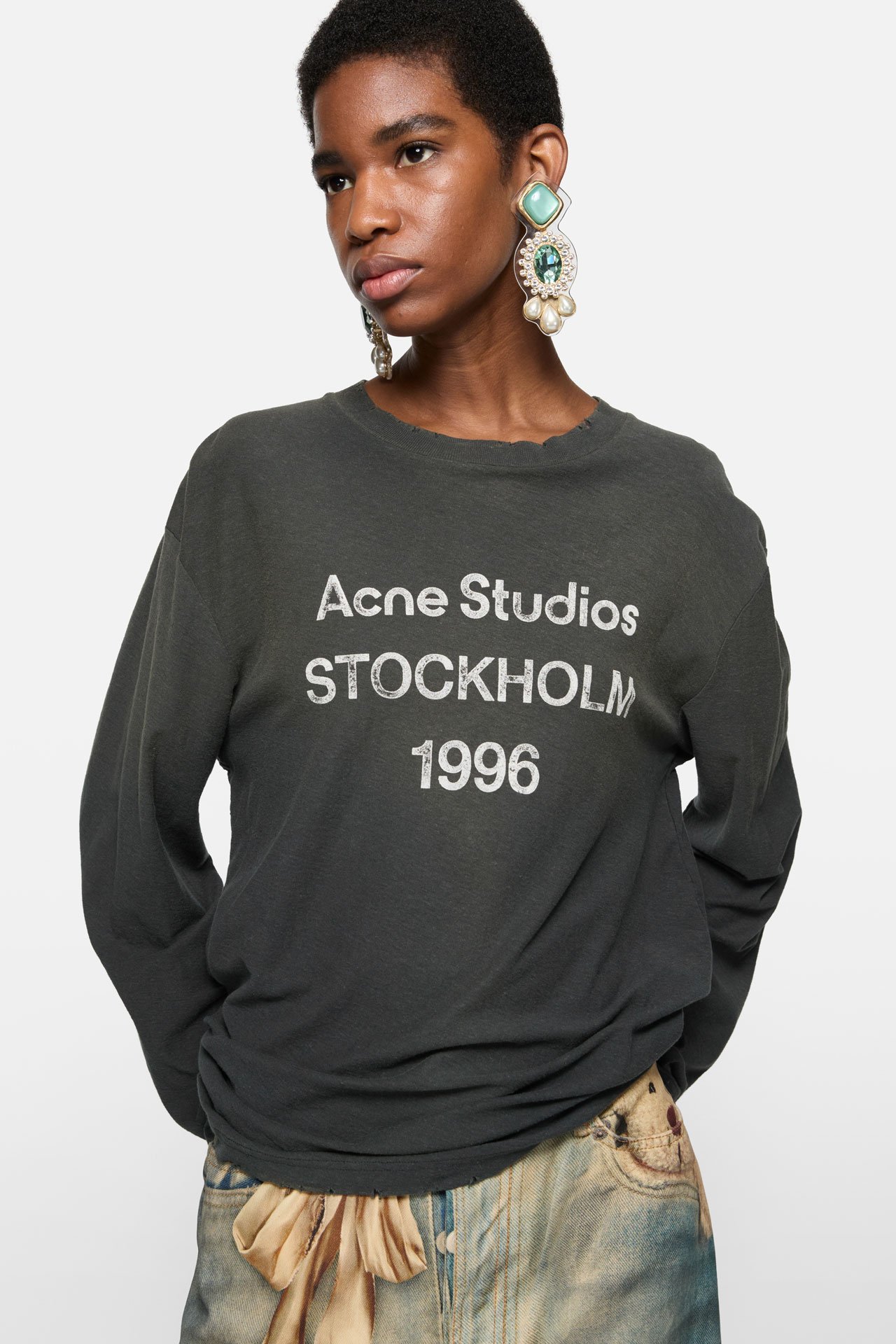 A short, black-haired woman wears bold blue earrings and black loose long-sleeve t-shirts by Acne Studios with the signature 1996 logo on it.