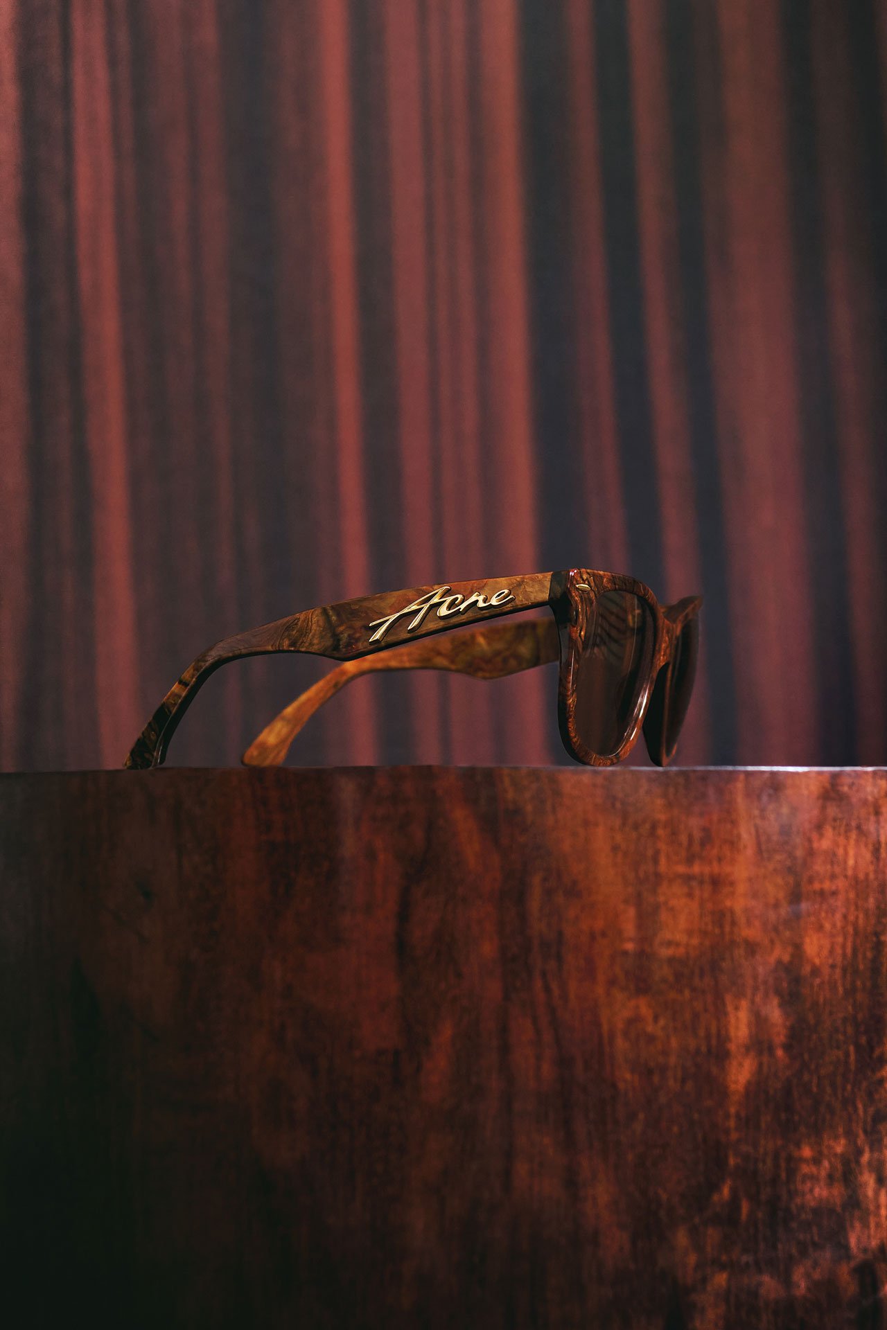 A showcase of the Acne Studios brown/gold square-frame sunglasses on a wooden pedestral.