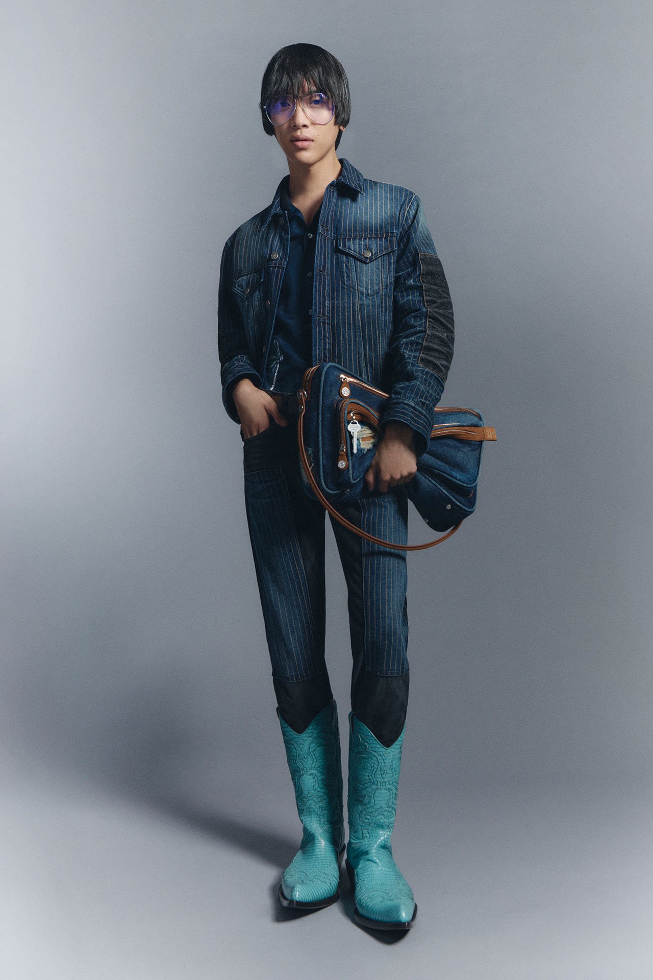 A male model wearing a matching blue, striped denim jacket and jeans, bright blue leather boots, holding a denim Camero bag.