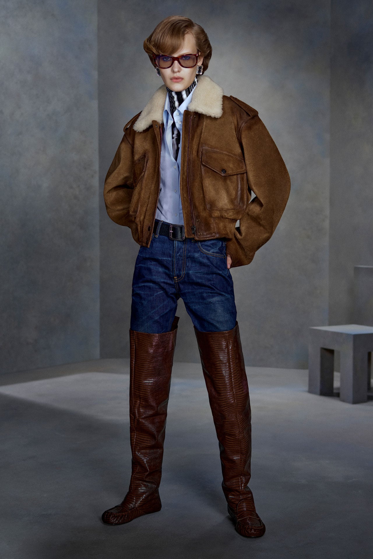 Acne Studios brown leather bomber jacket, dark blue jeans, light blue shirt and dark brown over-the-knee leather boots.