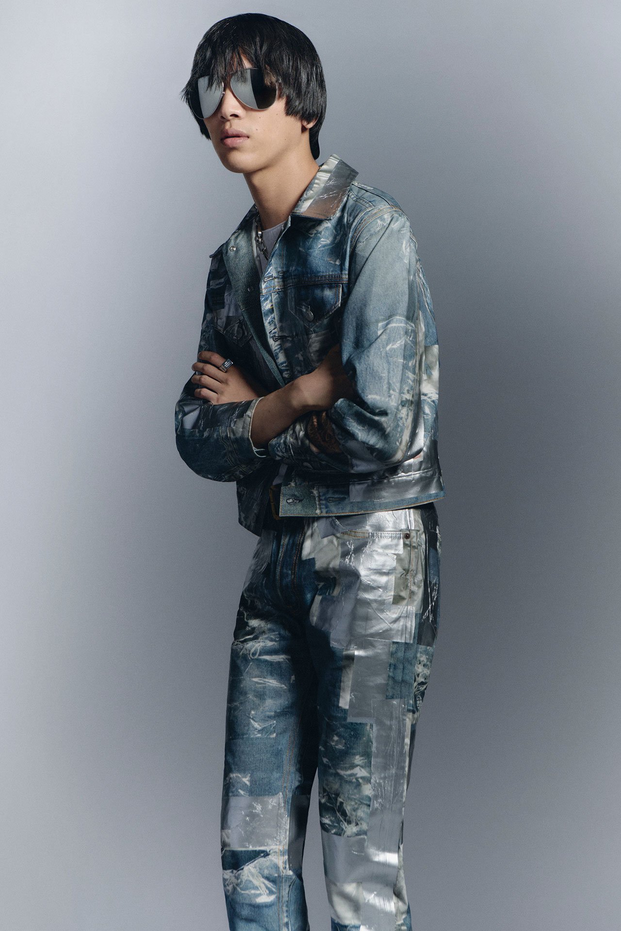 A close-up of a male model with glasses wearing Acne Studios SS26 new arrivals matching patchwork denim jacket and jeans.