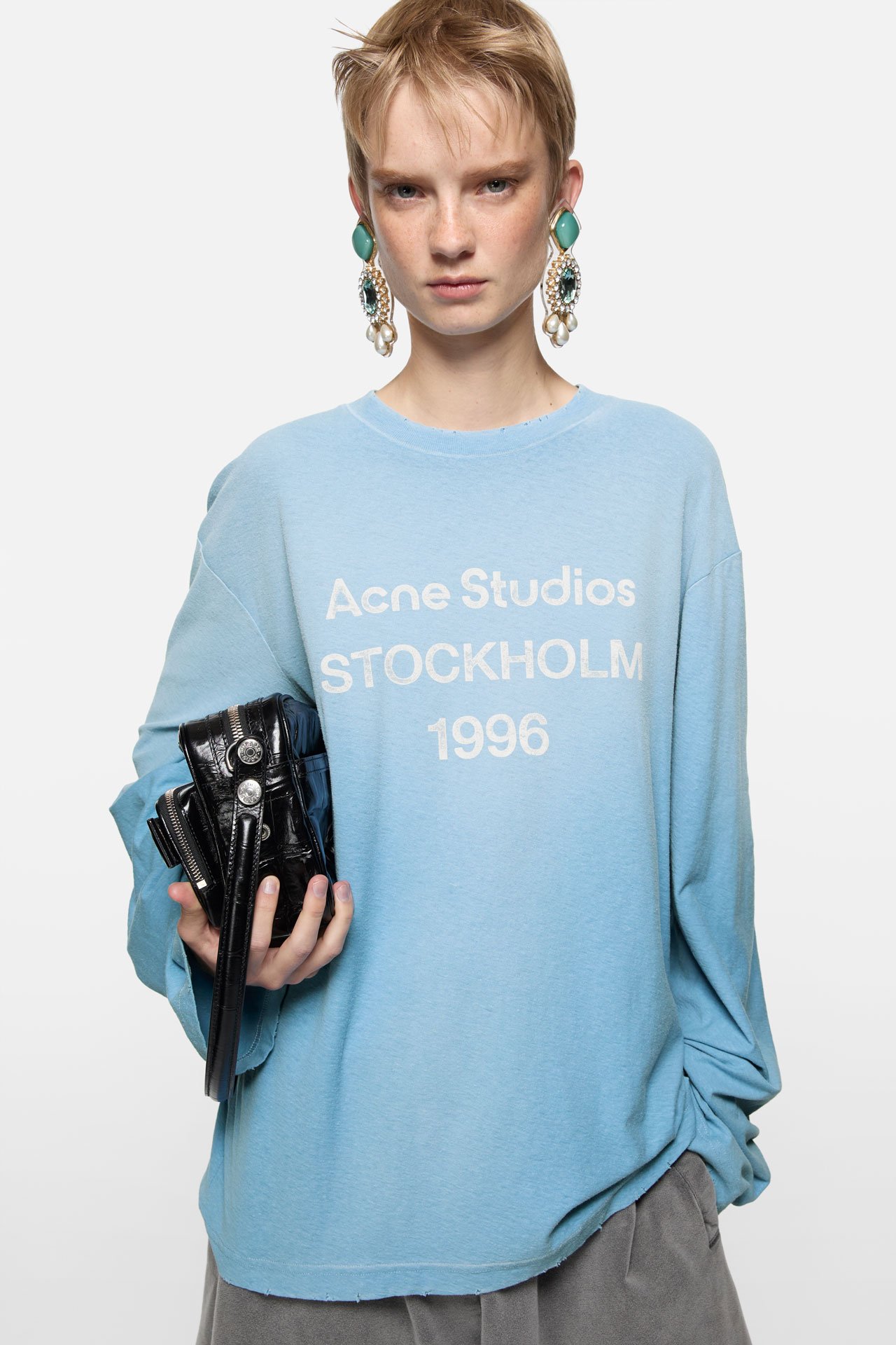 A female model wears bold blue earrings and cornflower blue loose long-sleeve t-shirts by Acne Studios with the signature 1996 logo on it, holding a black leather Camero bag.