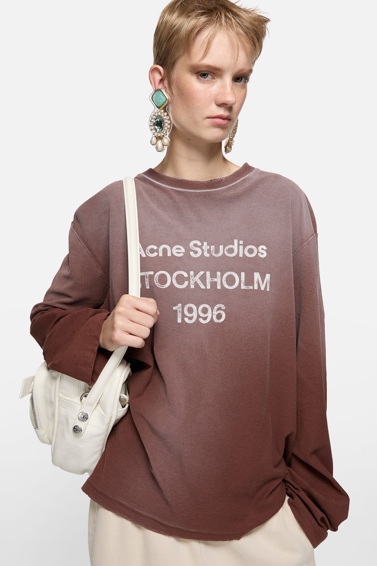 A blond short-haired female model wearing an Acne Studios 1996 logo brown long-sleeve shirt, a white Camero shoulder bag and teal earrings.