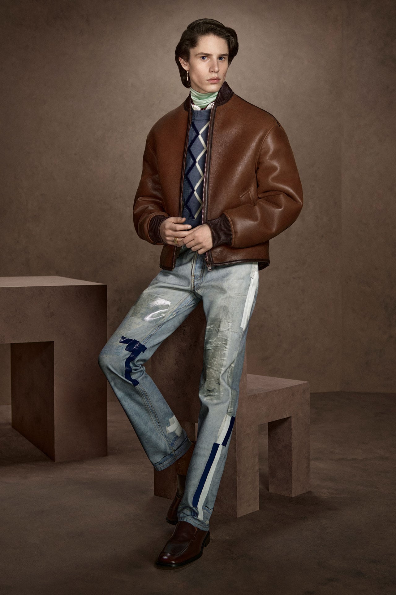 Acne Studios Menswear brown leather jacket, blue sweater, blue TLO tape jeans and brown leather loafers.