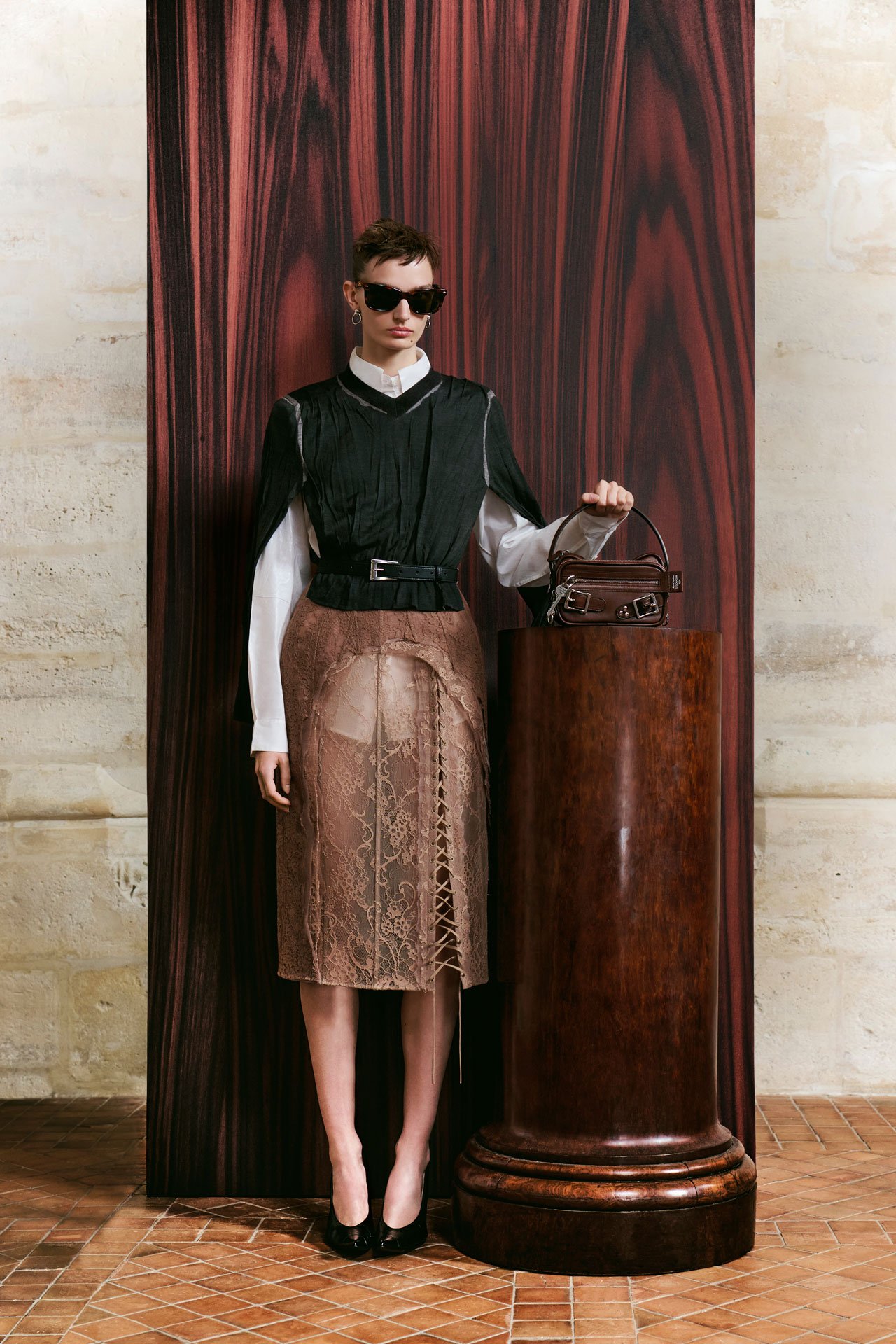 Acne Studios SS26 Runway - a female model wearing a black knitted top with a white long-arm top under it, taupe lace-skirt and black sunglasses, holding a small brown Camero bag.