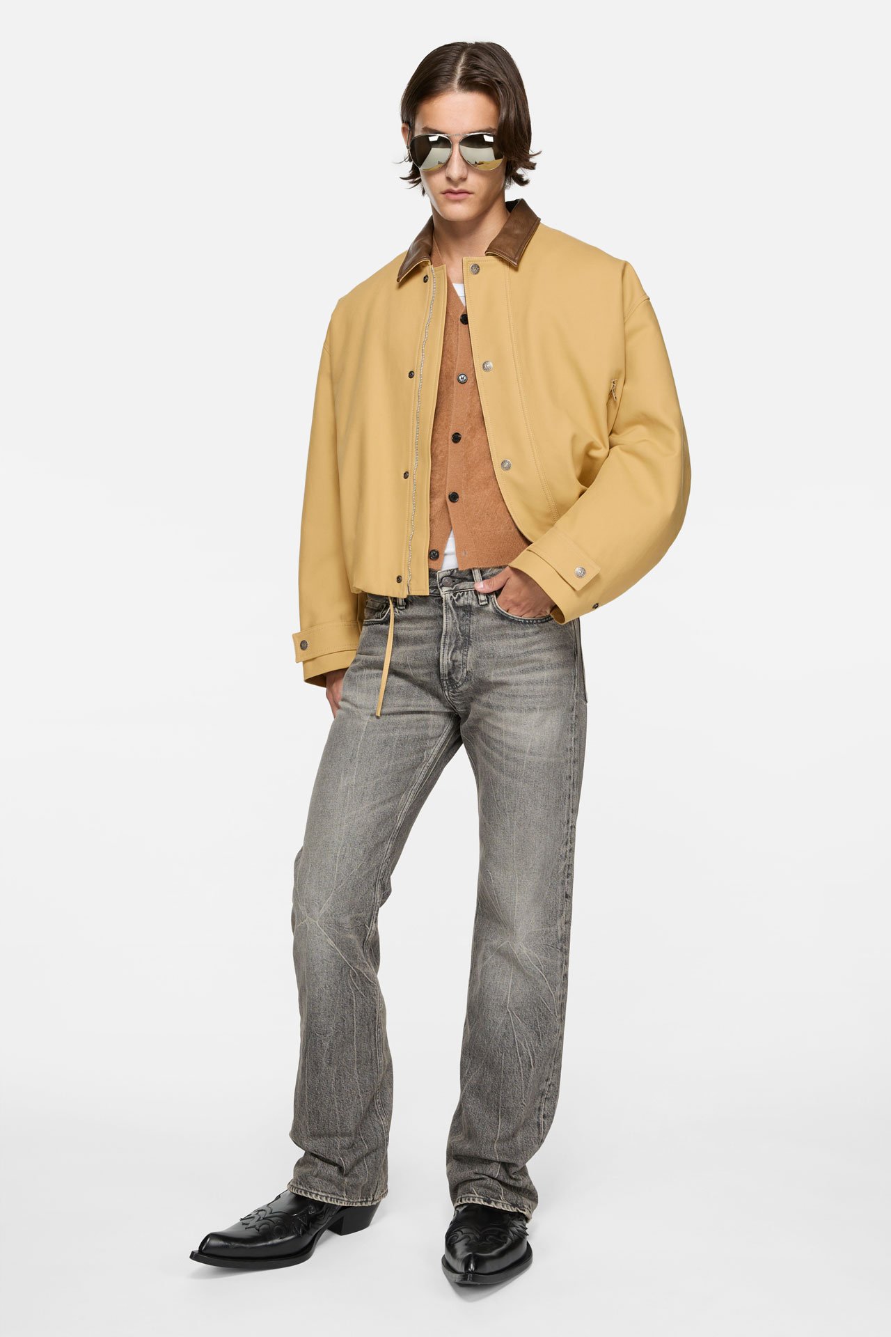 A male model wearing an Acne Studios grey regular fit jeans in 2010M style, a beige bomber jacket with leather details and mirror glasses.