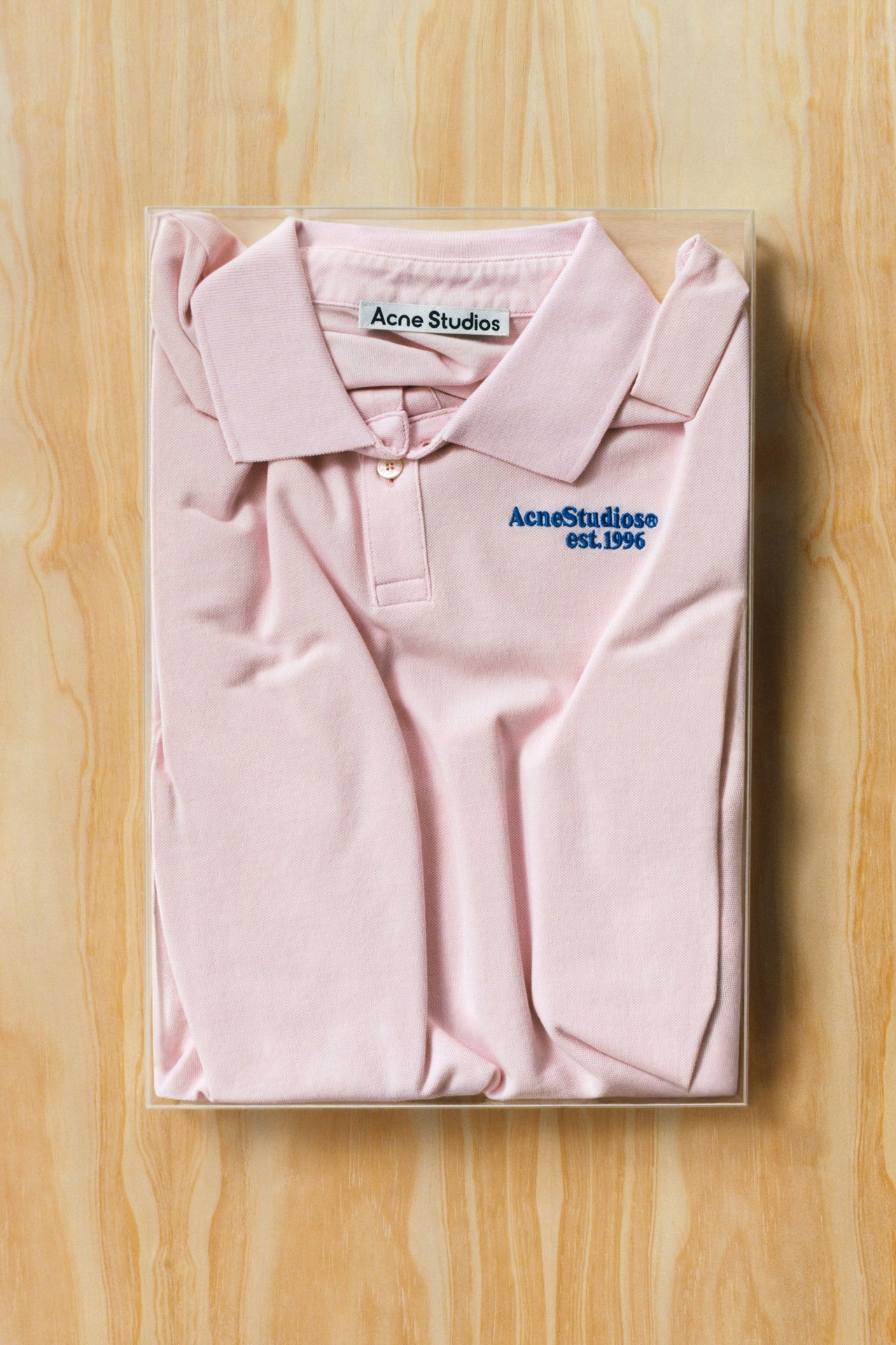 Acne Studios SS26 Summer Collection light pink t-shirt with collar and blue Acne Studios logo folded and presented on a light wooden tray.