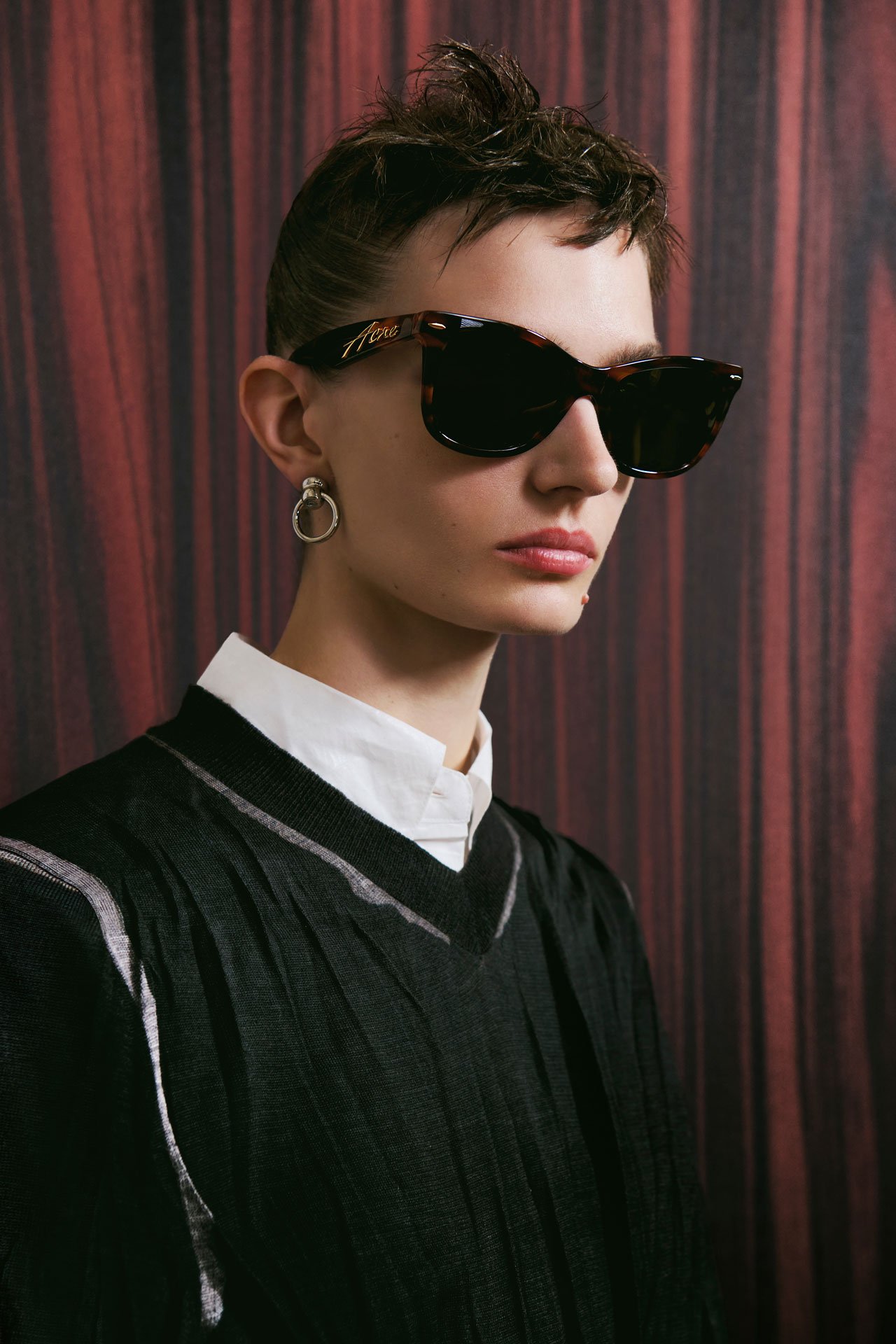 Female model wearing Acne Studios brown/gold square-frame sunglasses and a sheer black knitted shirt and gold earrings.