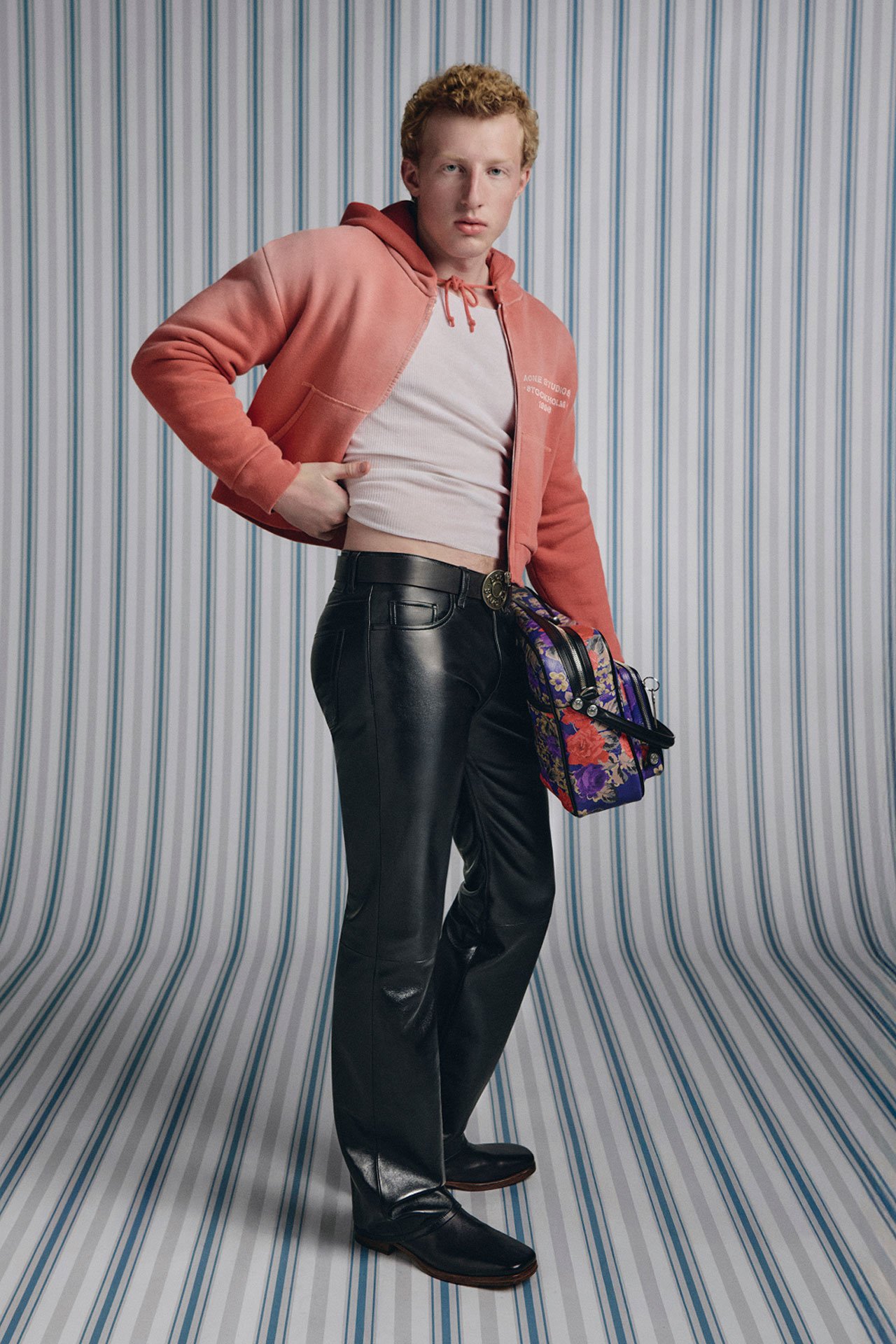 SS26 Acne Studios Mens Sunkissed - male model wearing a red hoodie, white tanktop, black leather pants and holding a purple flower-printed leather Camero bag.
