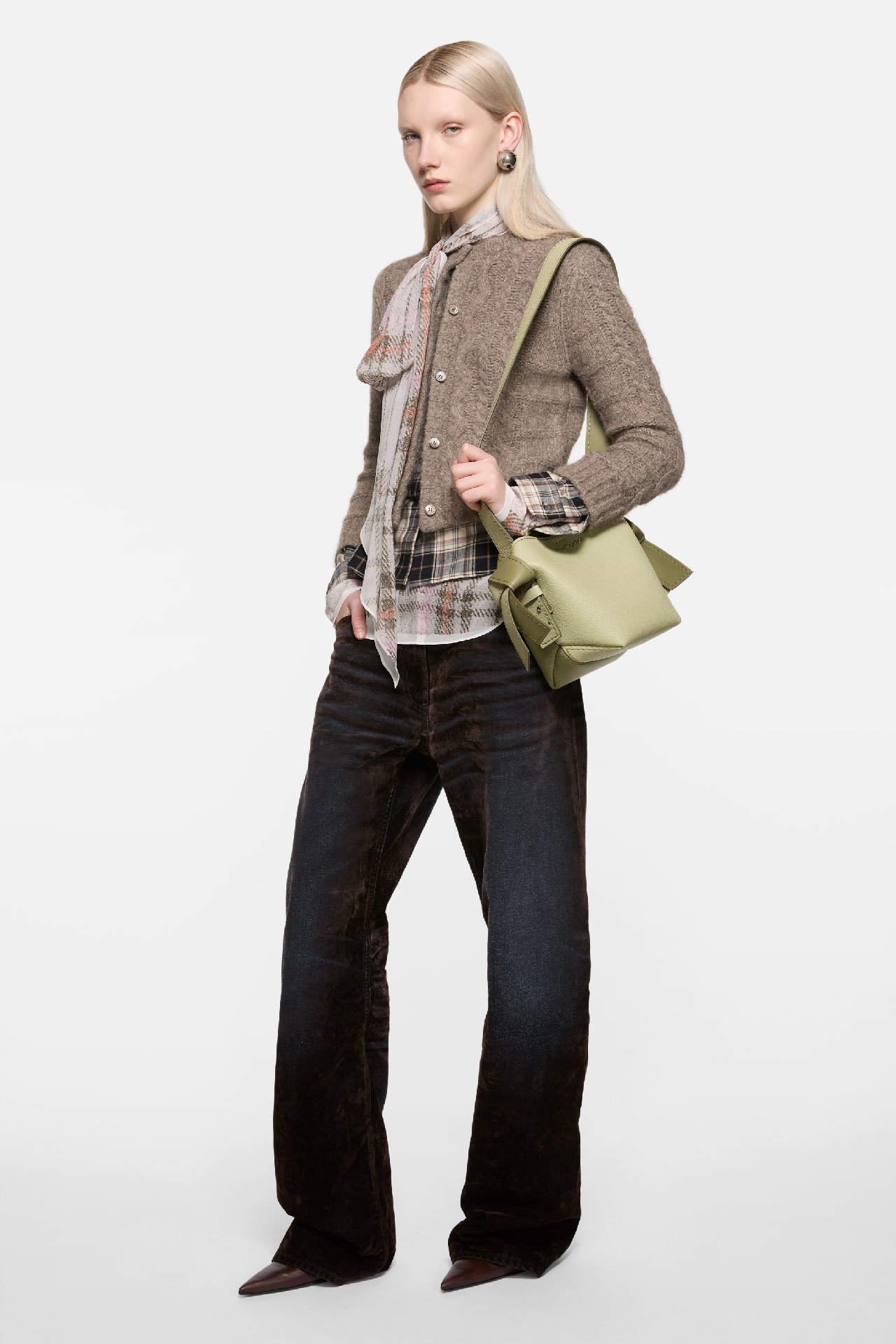 Female model wearing an Acne Studios Musubi mini shoulder bag in sage green, beige knitted cardigan, dark blue jeans and soft silhouettes.
