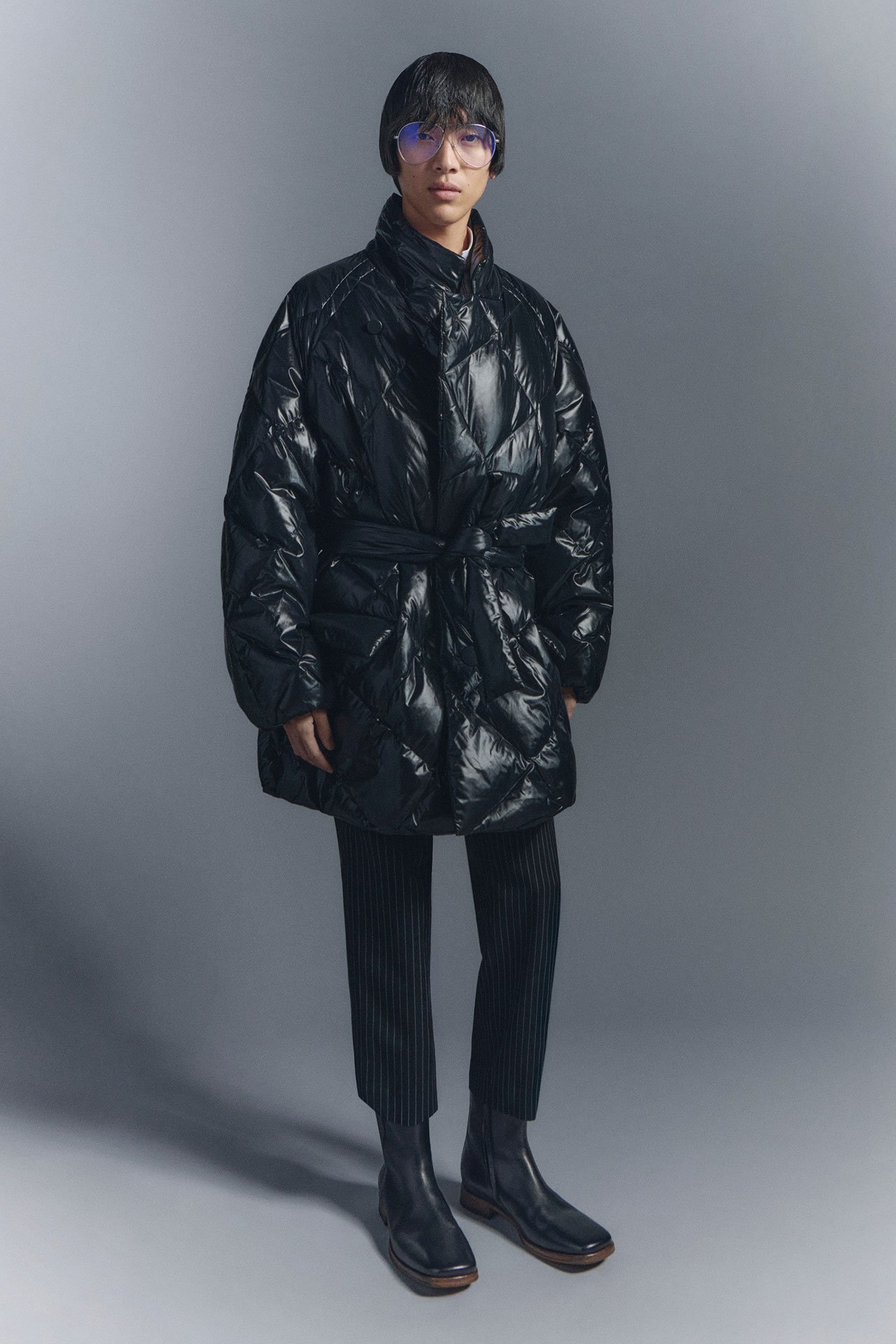 A black haired man in Acne Studios SS26 new arrivals black belted winter coat, black striped tailored pants and black leather boots.