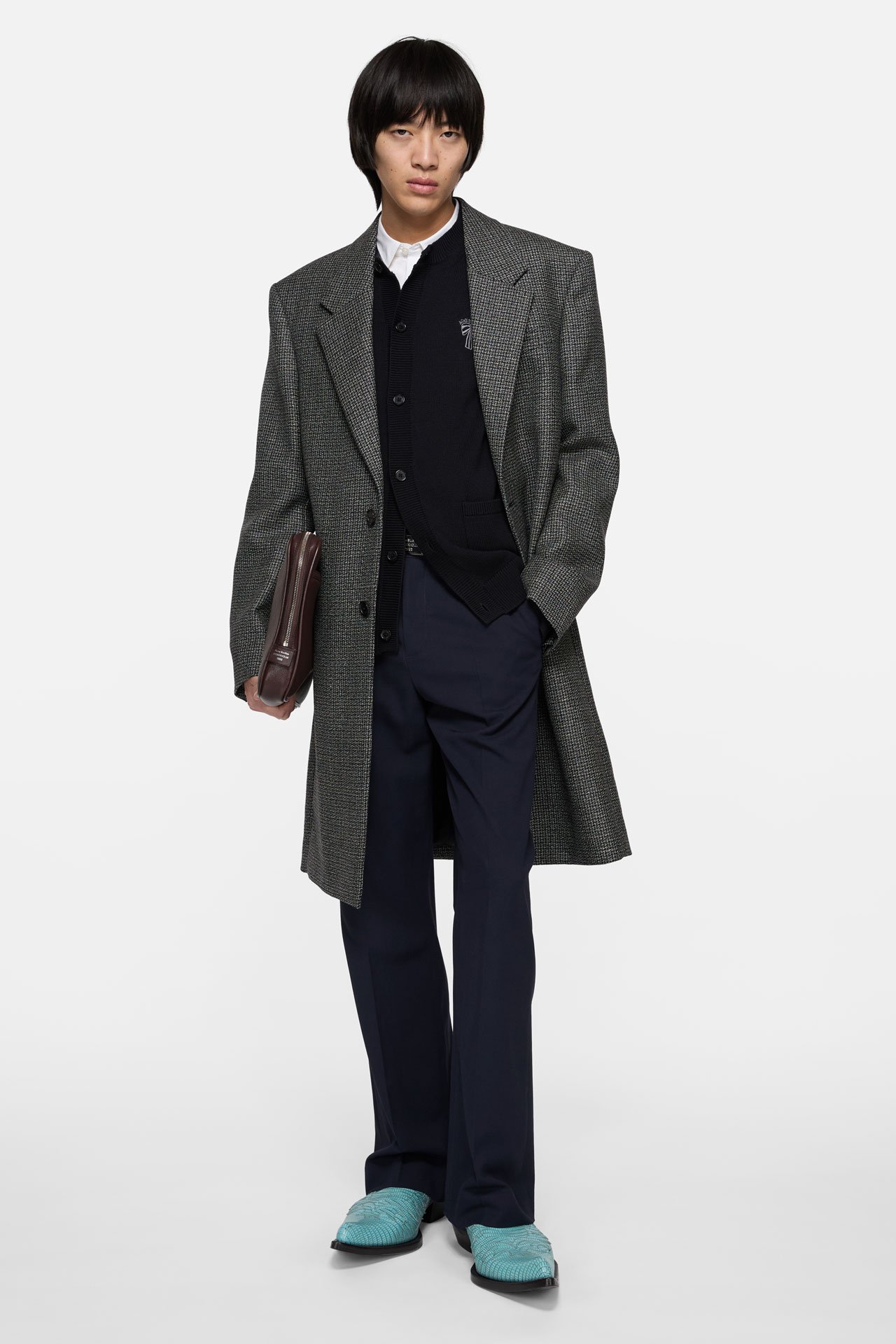 A male model wearing Acne Studios blue tailored trousers, black knitted cardigan, grey checked tailored suit jacket, blue leather cowboy boots and holding a brown leather laptop case.